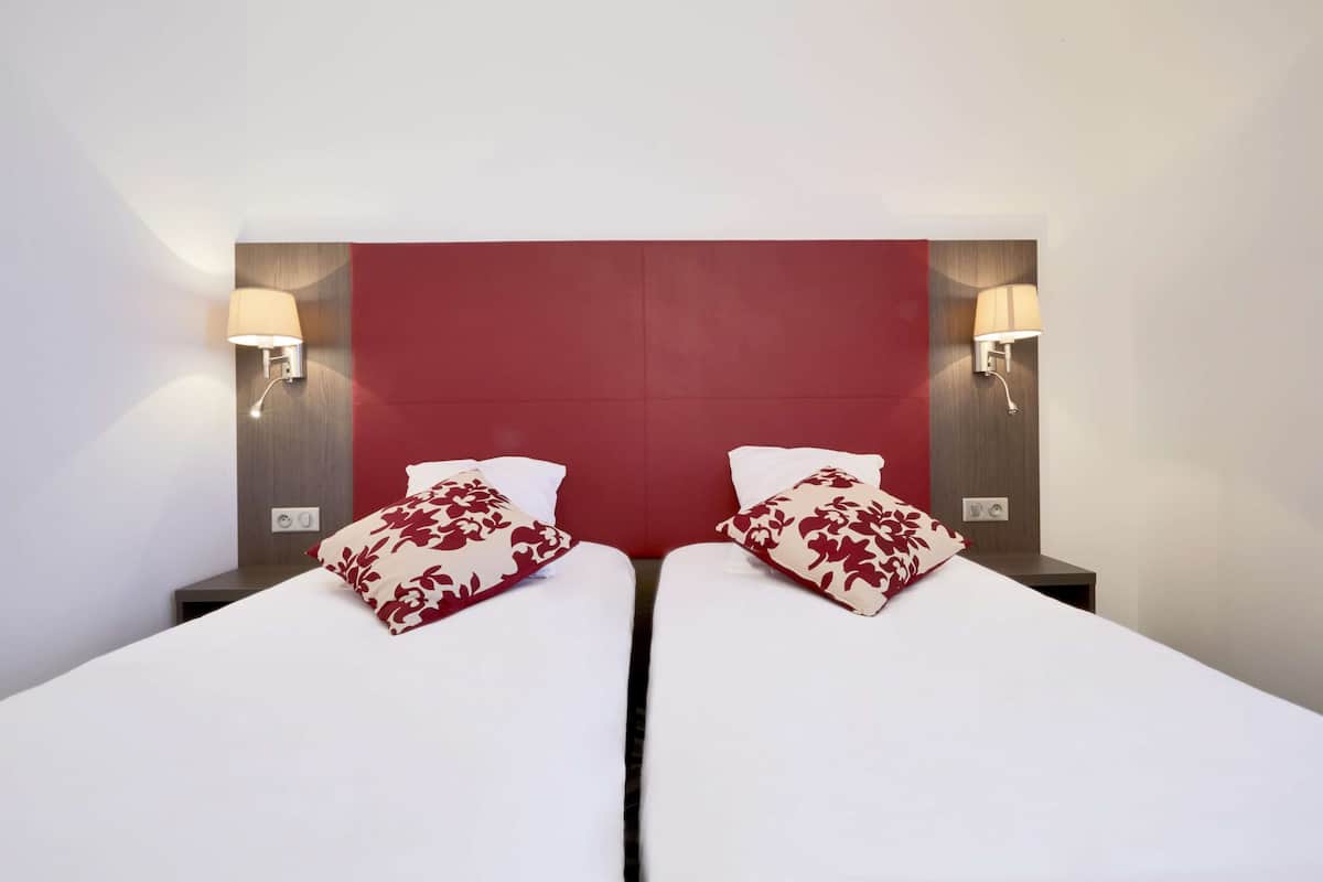 Ein Hotelzimmer mit zwei Betten, jeweils mit weißer Bettwäsche und roten Kissen mit Blumenmuster. An beiden Seiten der Betten befinden sich Wandlampen.