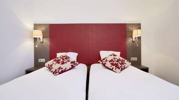 A hotel room with two beds, each with white bedding and red floral pillows. There are wall-mounted lamps on either side of the beds.