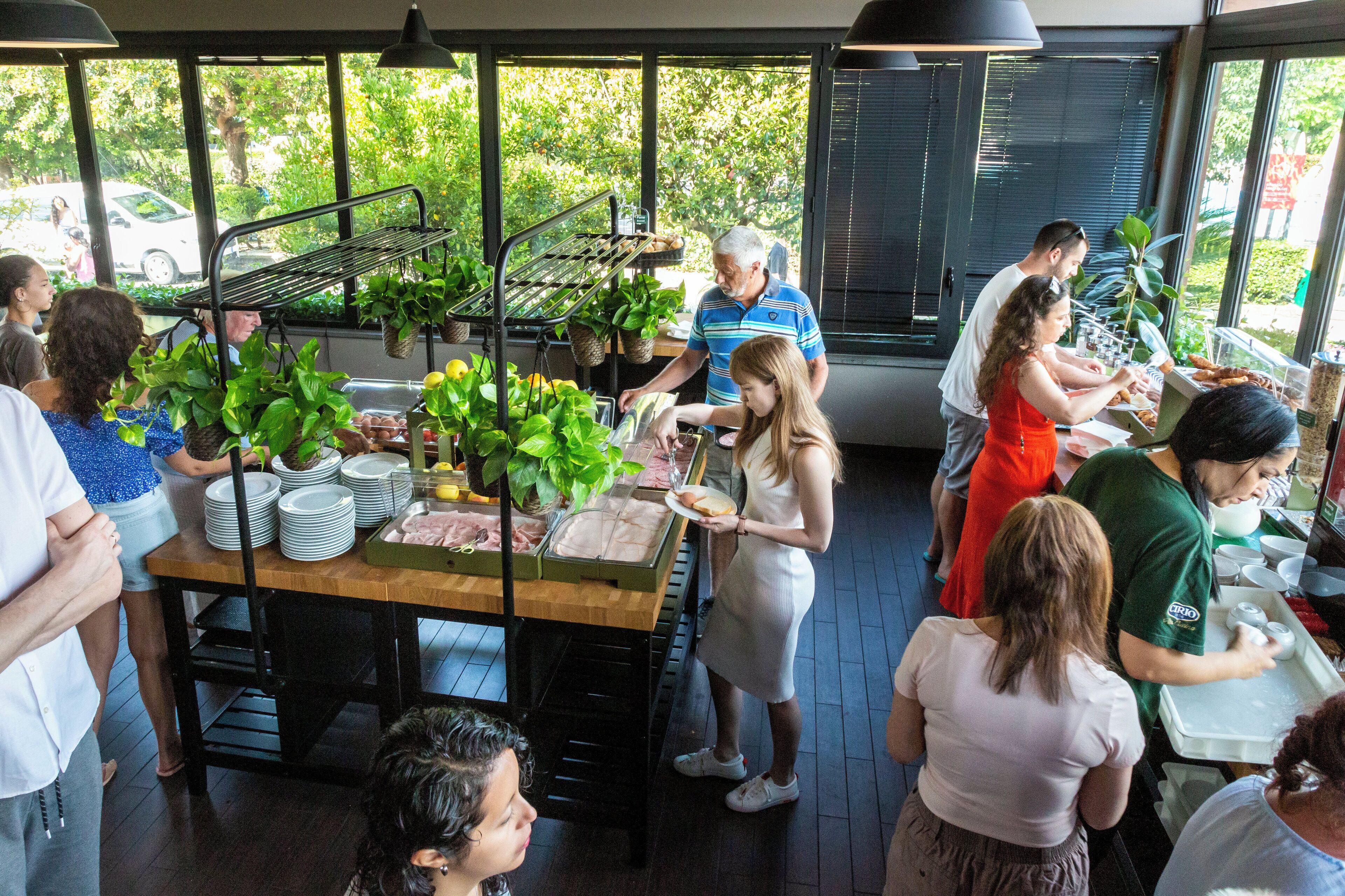 A group of people in a restaurant with a large window, a wooden table with plates and food, and a hanging plant rack.