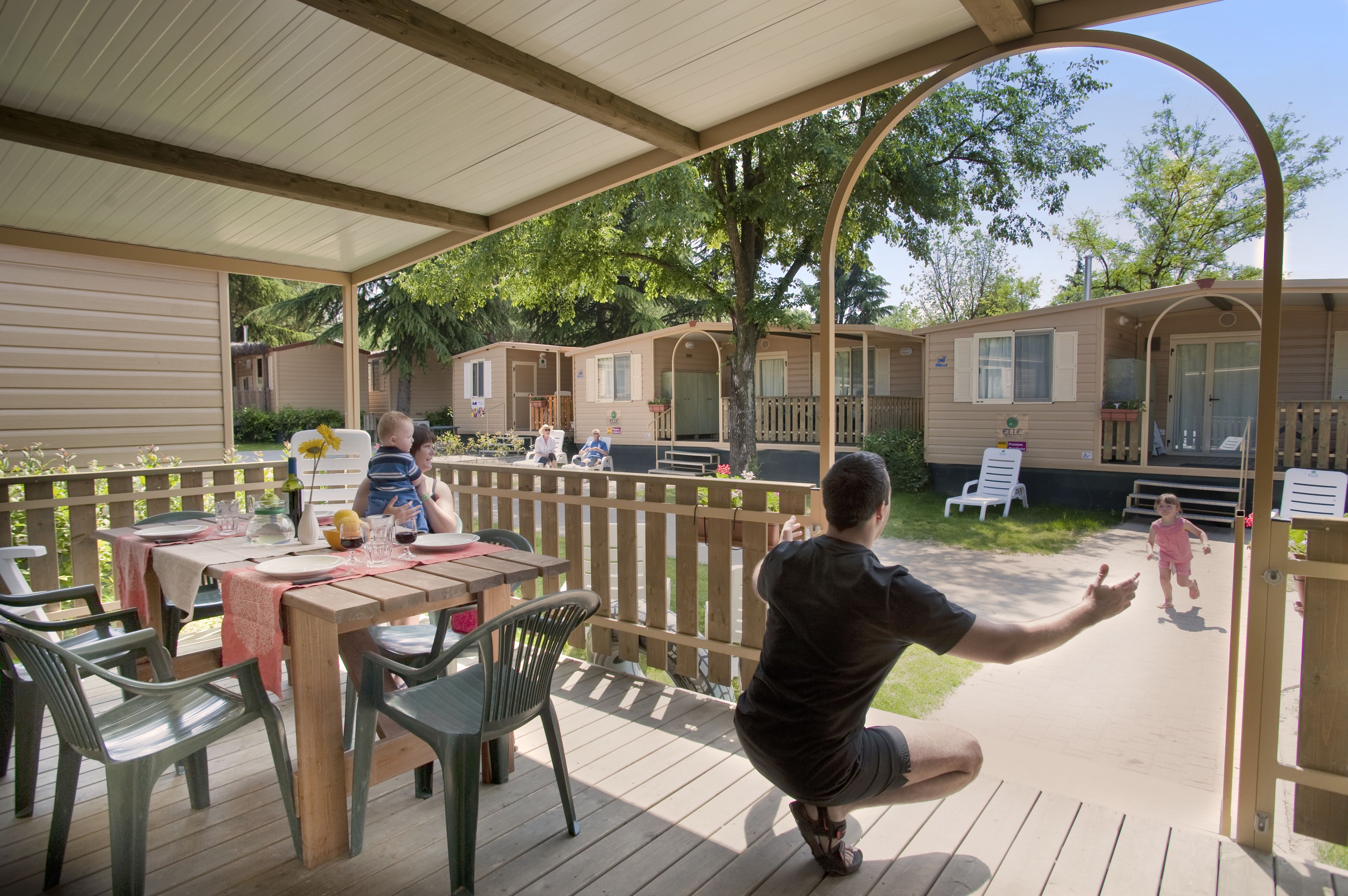 Un espacio al aire libre cubierto con mesas y sillas de picnic, una terraza de madera y vista a casas móviles al fondo.