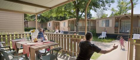 A covered outdoor area with picnic tables and chairs, a wooden deck, and a view of mobile homes in the background.