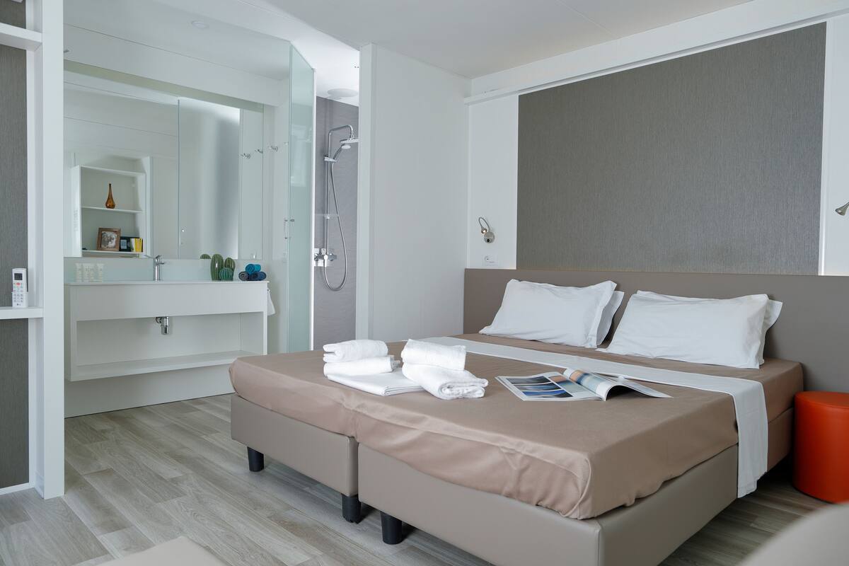 Moderni hotellihuone, jossa on suuri sänky, seinään upotettu suihku, peilillä varustettu meikkipöytä ja pieni oranssi jakkara.