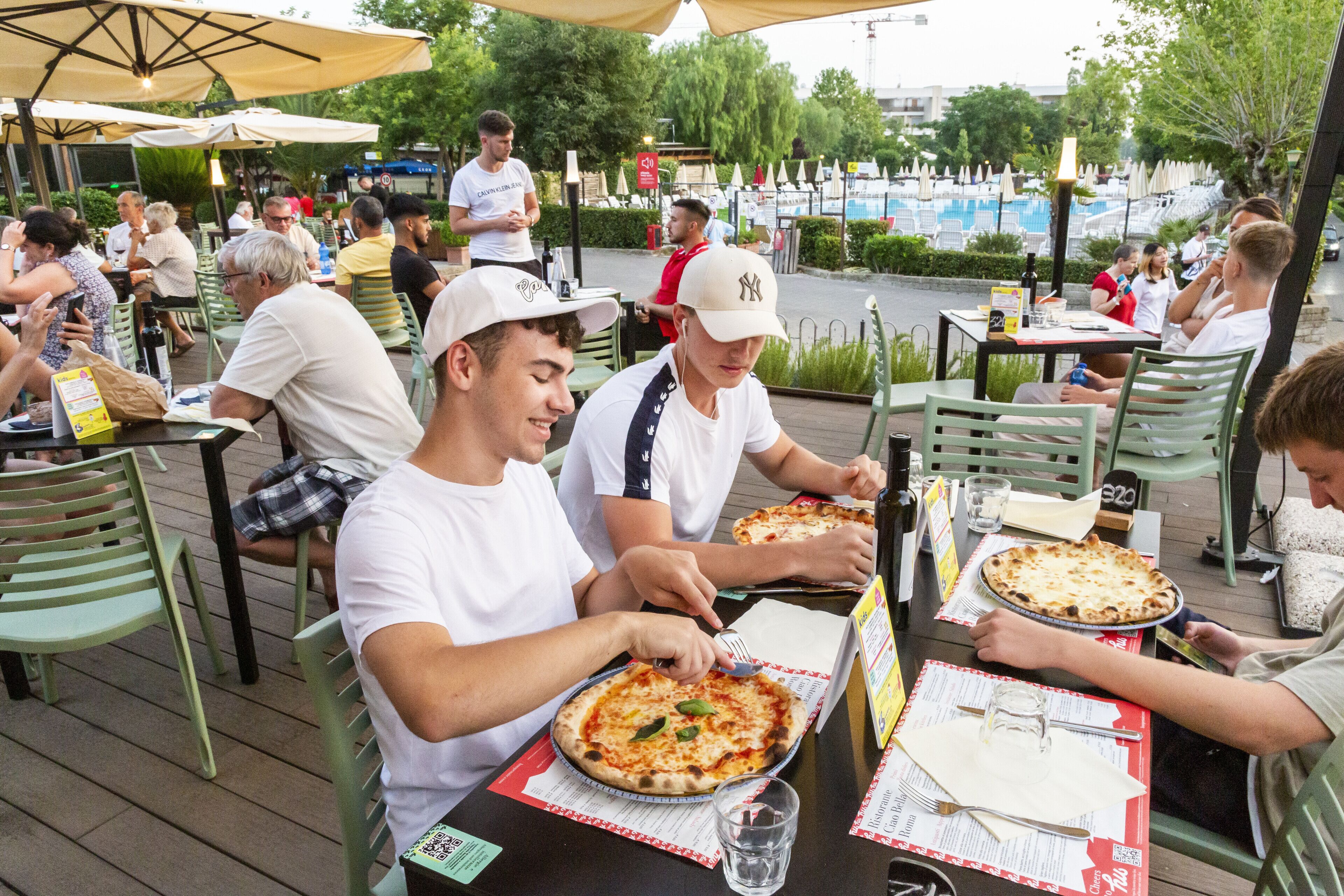 An outdoor dining area with tables and chairs, a pool in the background, and people eating.