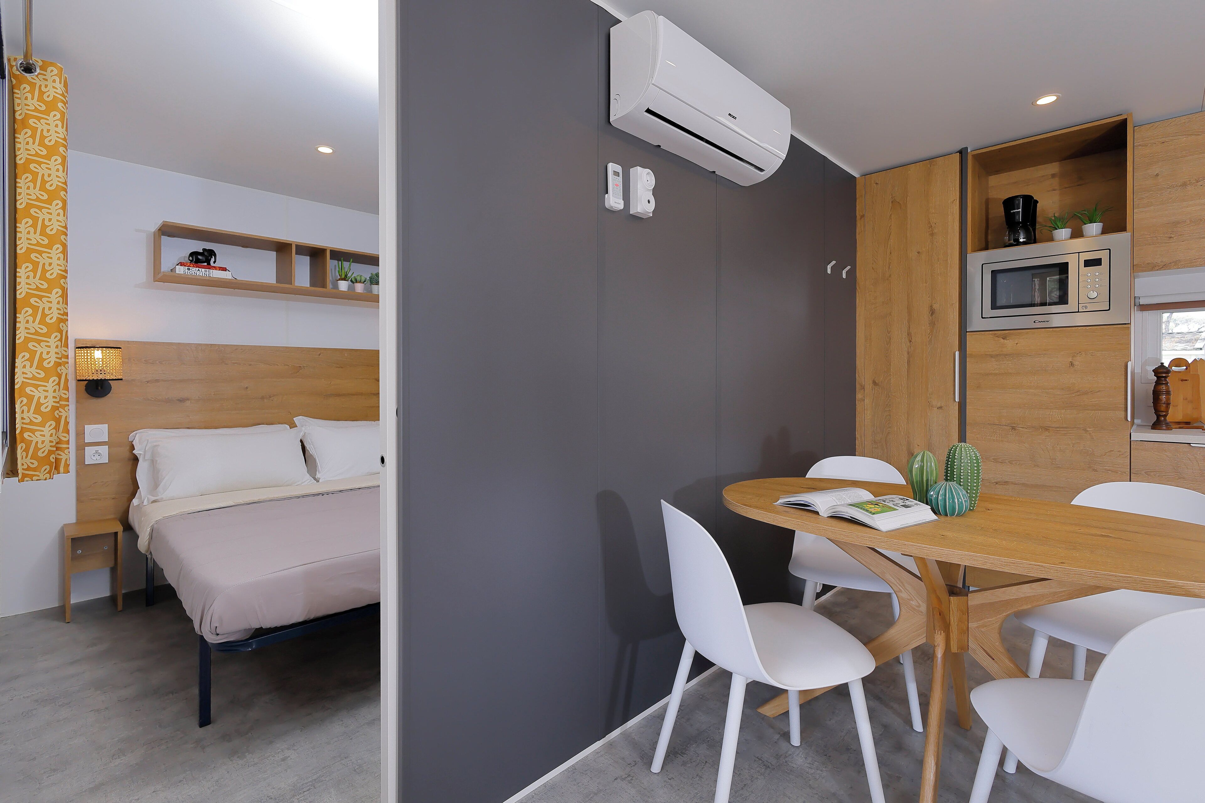 A modern hotel room with a bed, a dining table with chairs, a microwave, and an air conditioning unit.