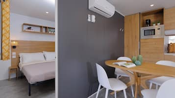 A modern hotel room with a bed, a dining table with chairs, a microwave, and an air conditioning unit.