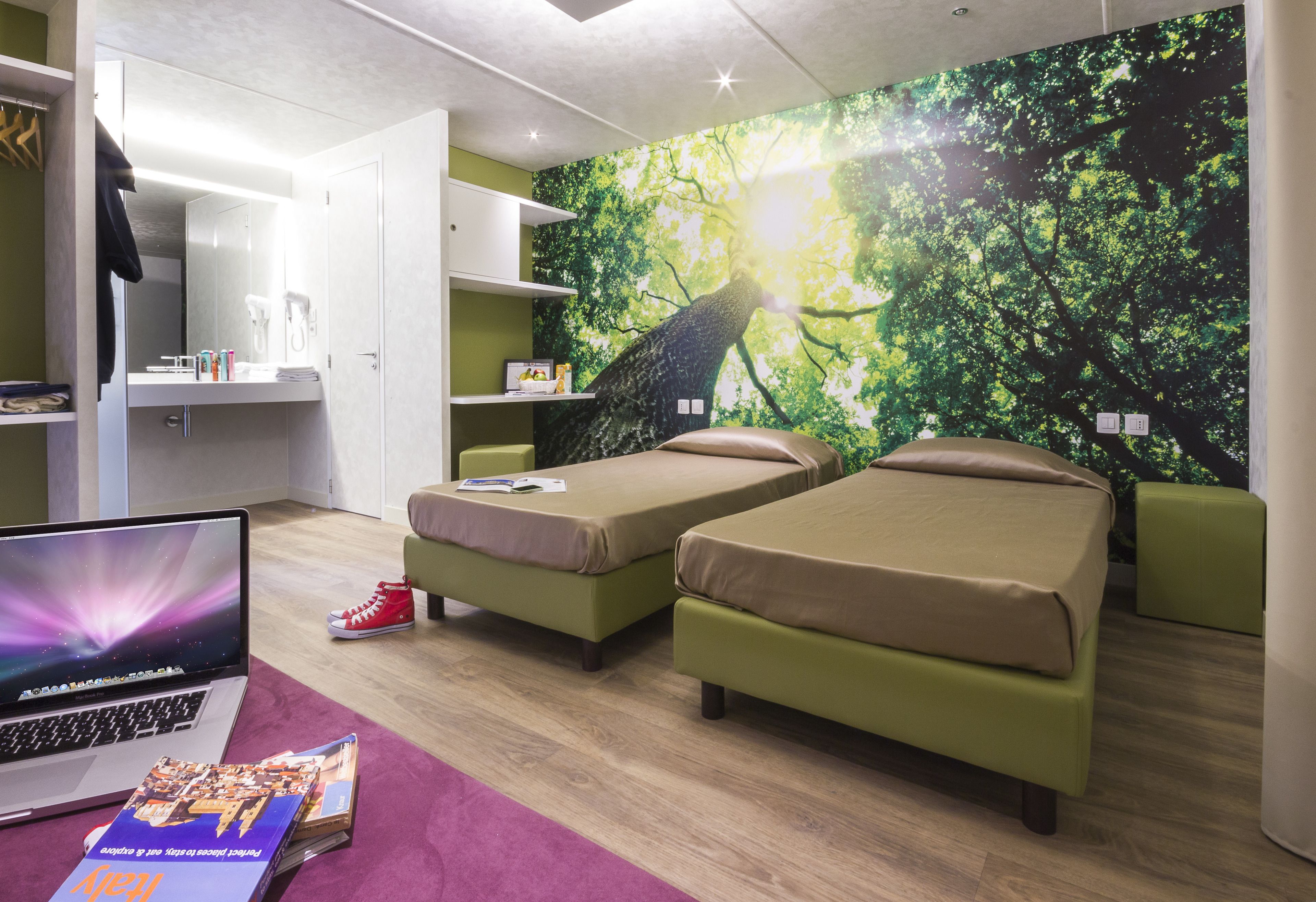 A modern bedroom with two single beds, a desk with a laptop, and a large tree mural on the wall.