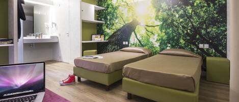 A modern bedroom with two single beds, a desk with a laptop, and a large tree mural on the wall.