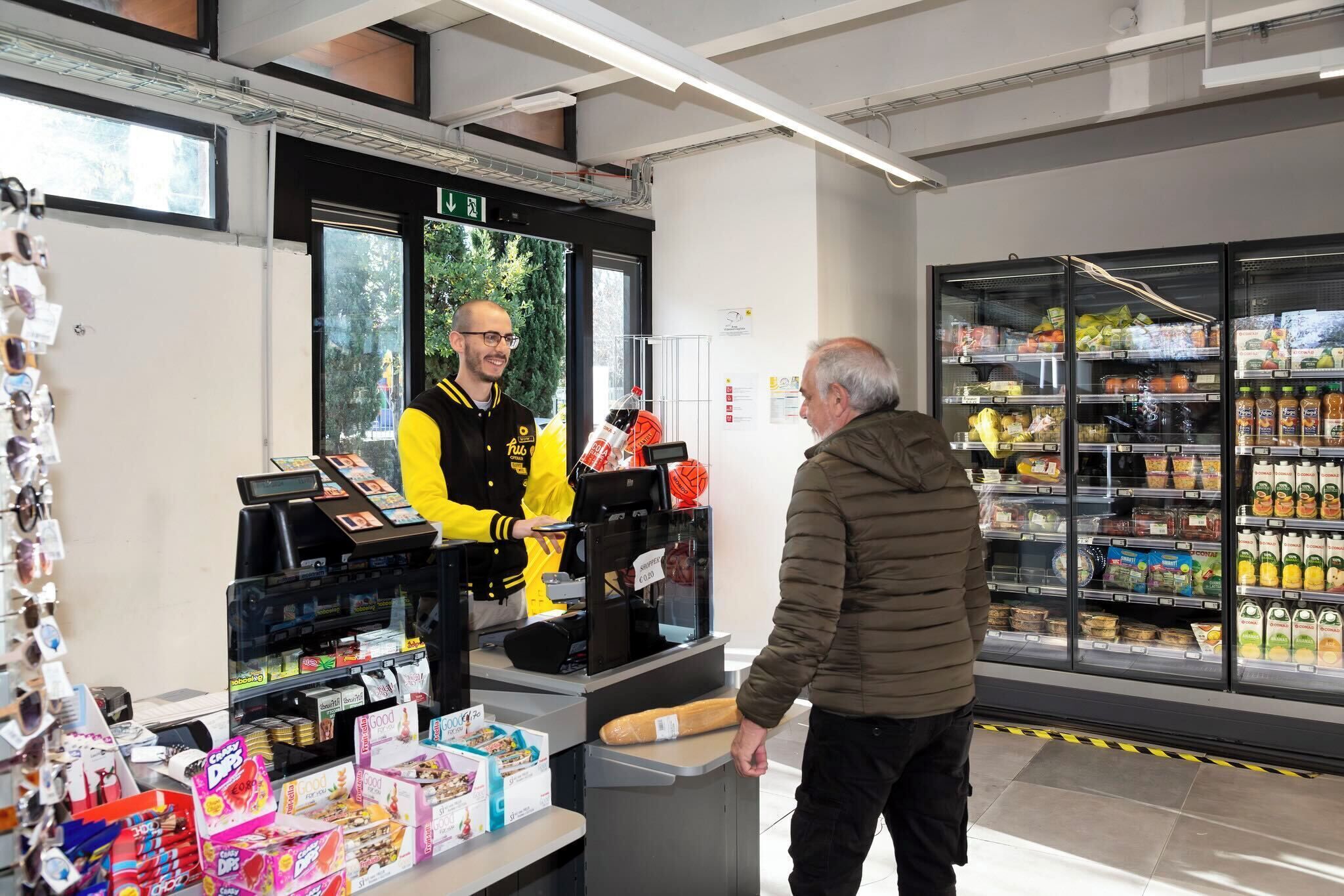 Un área de autoservicio con una terminal de pago digital, un refrigerador con diversos productos y un mostrador con artículos envasados.