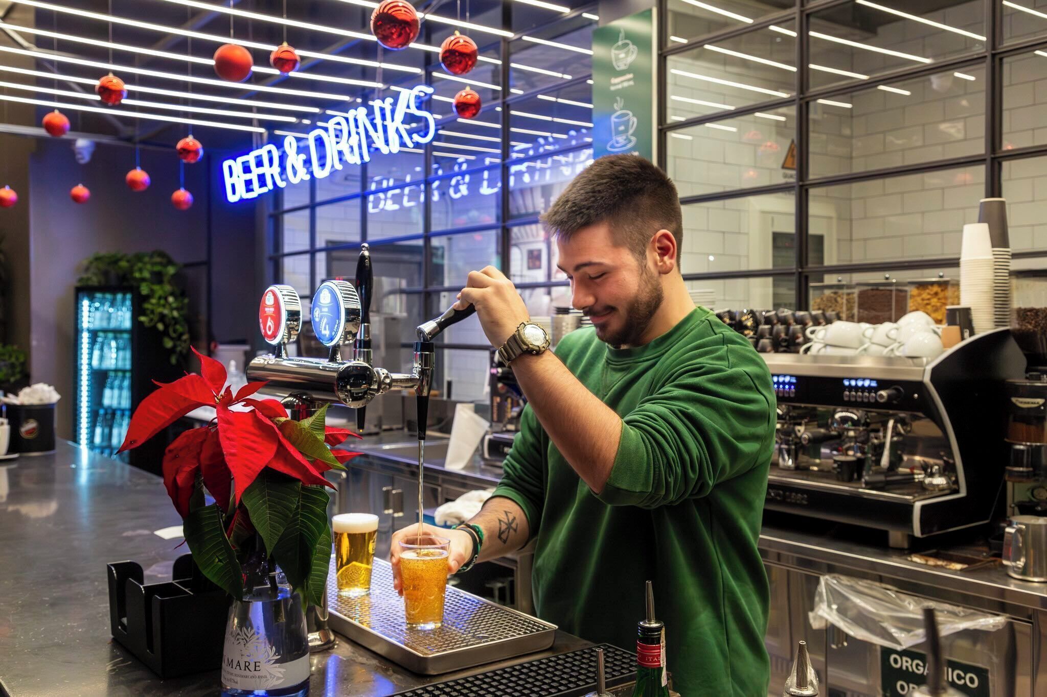 Een barista schenkt bier in een glas aan een bar met een biertap en een neonreclame op de achtergrond.