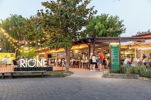 An outdoor seating area with tables and chairs, a sign reading "RIONE", and a tree in the background.