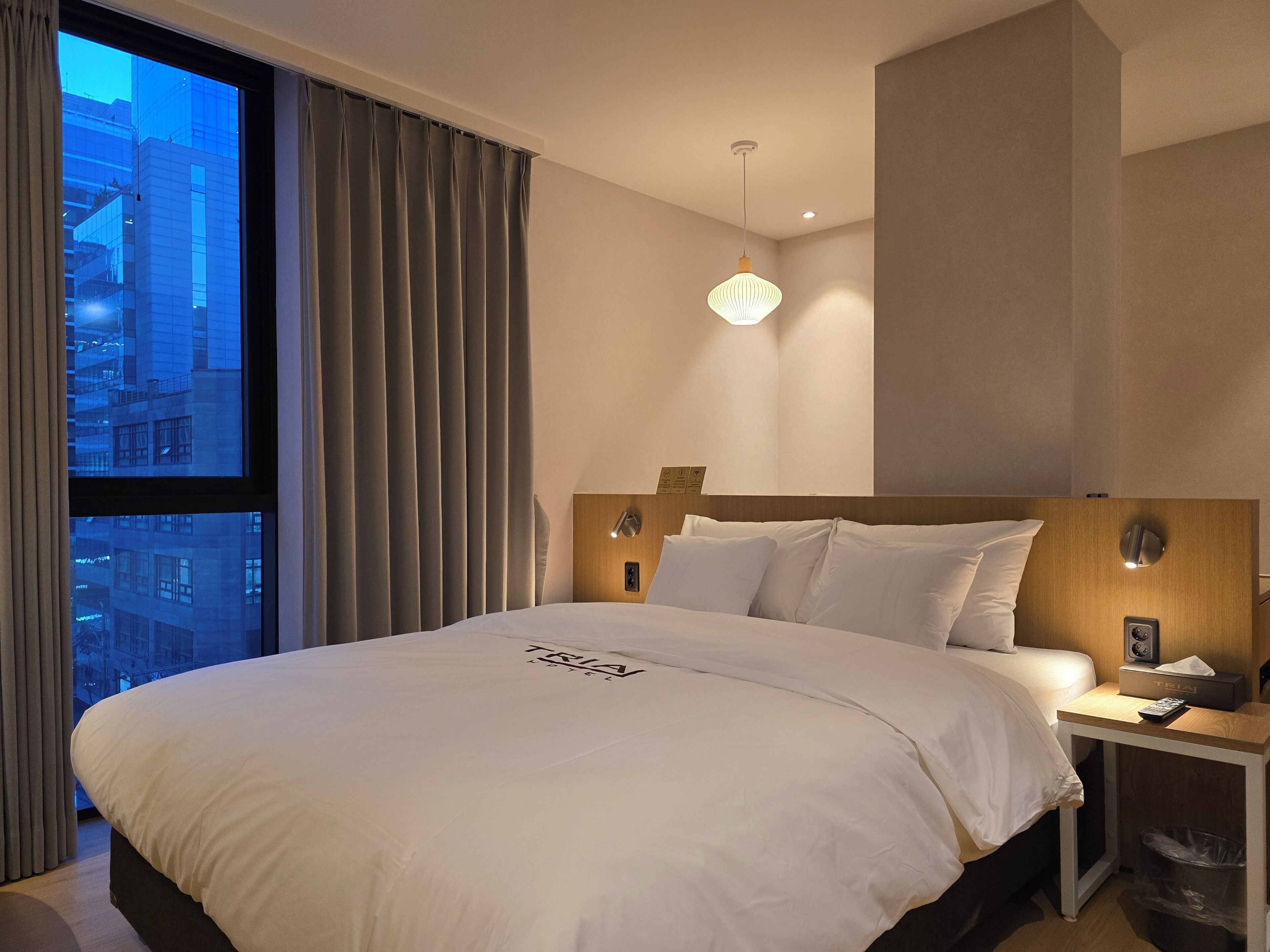 A hotel room with a large bed, a nightstand, a wall-mounted lamp, and a window with curtains.