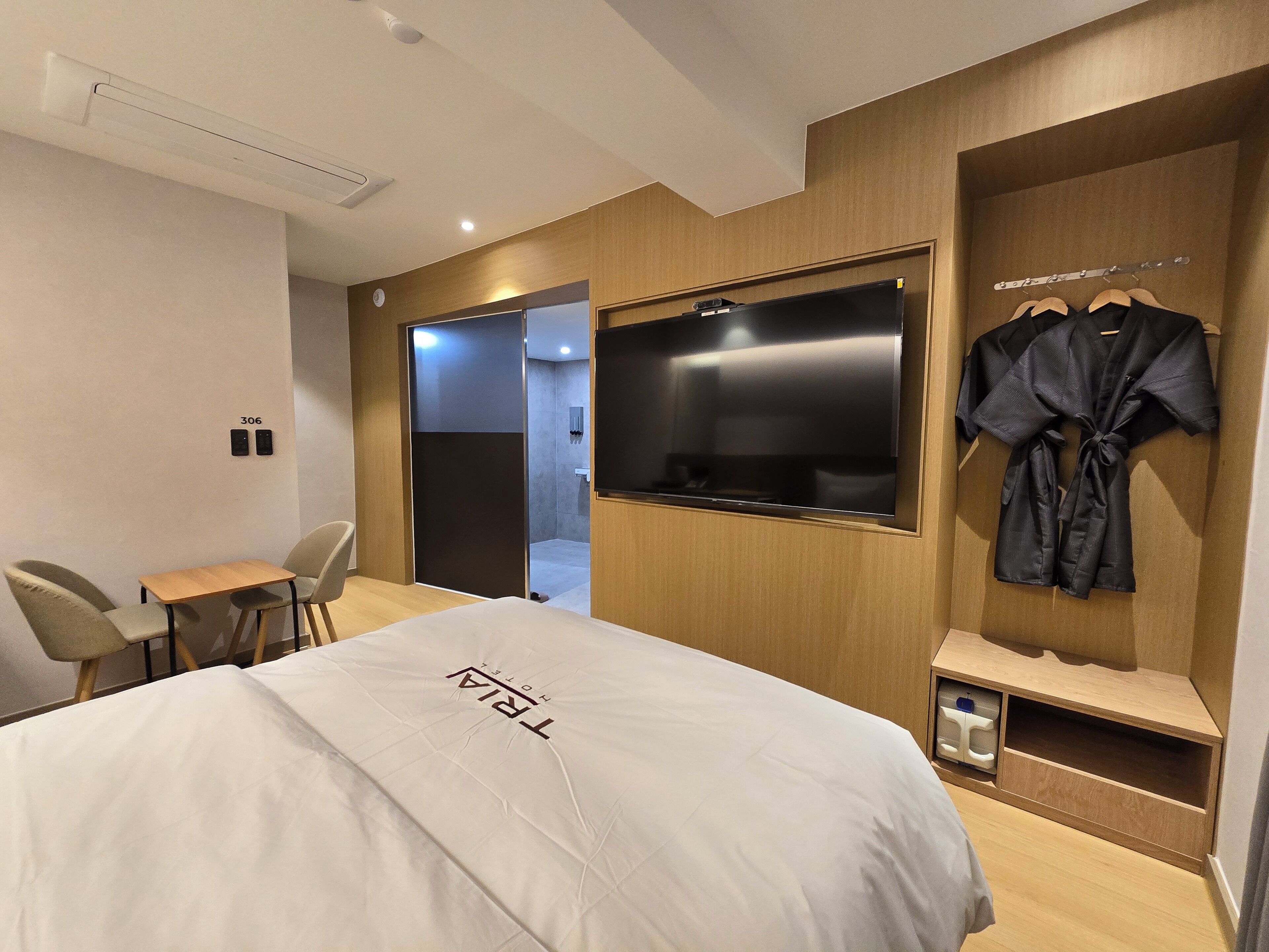 A hotel room with a bed, a TV mounted on the wall, a robe hanging on a rack, and a small table with chairs.