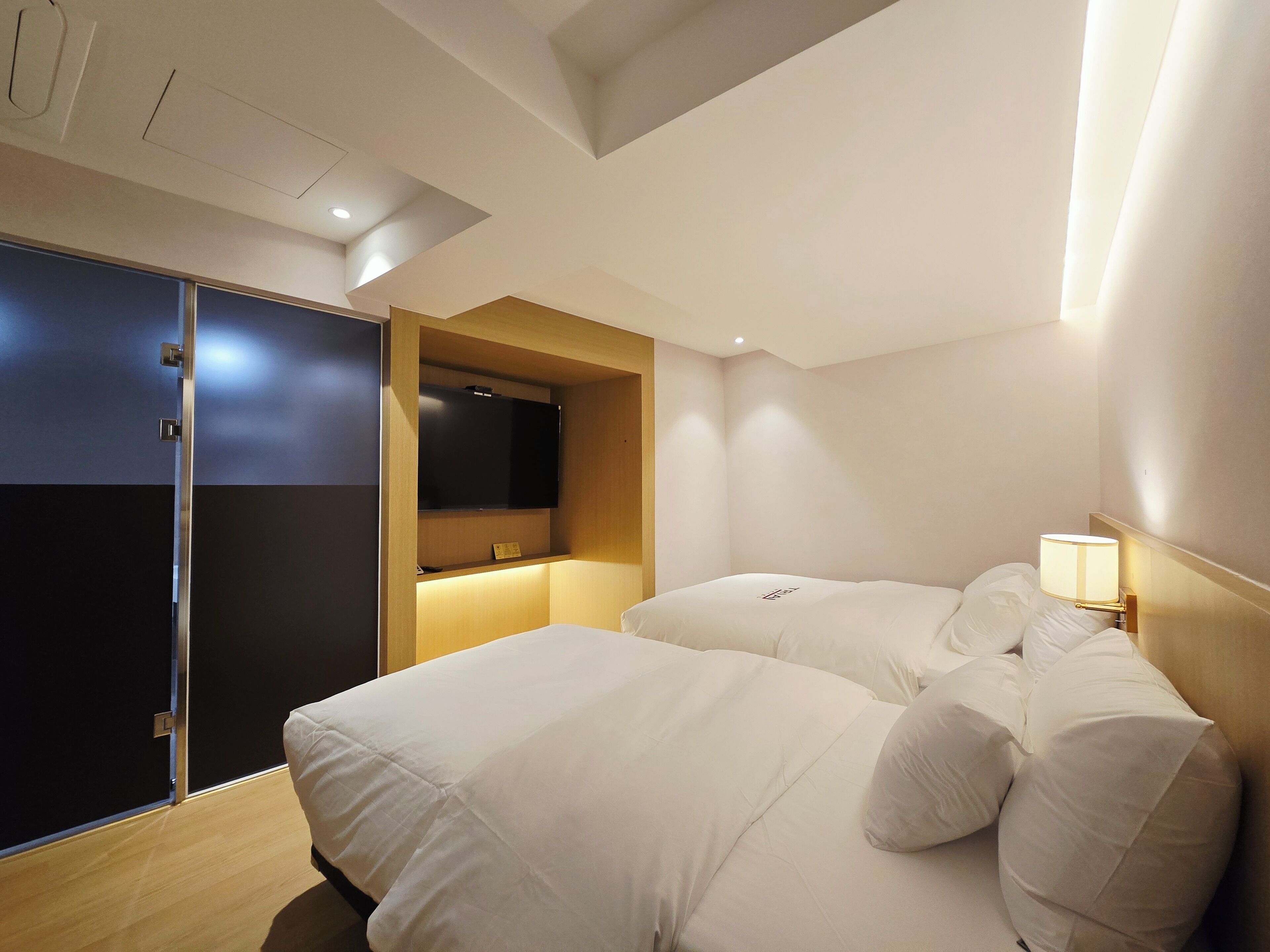 A hotel room with two beds, a TV mounted on a wooden panel, and a bedside lamp.
