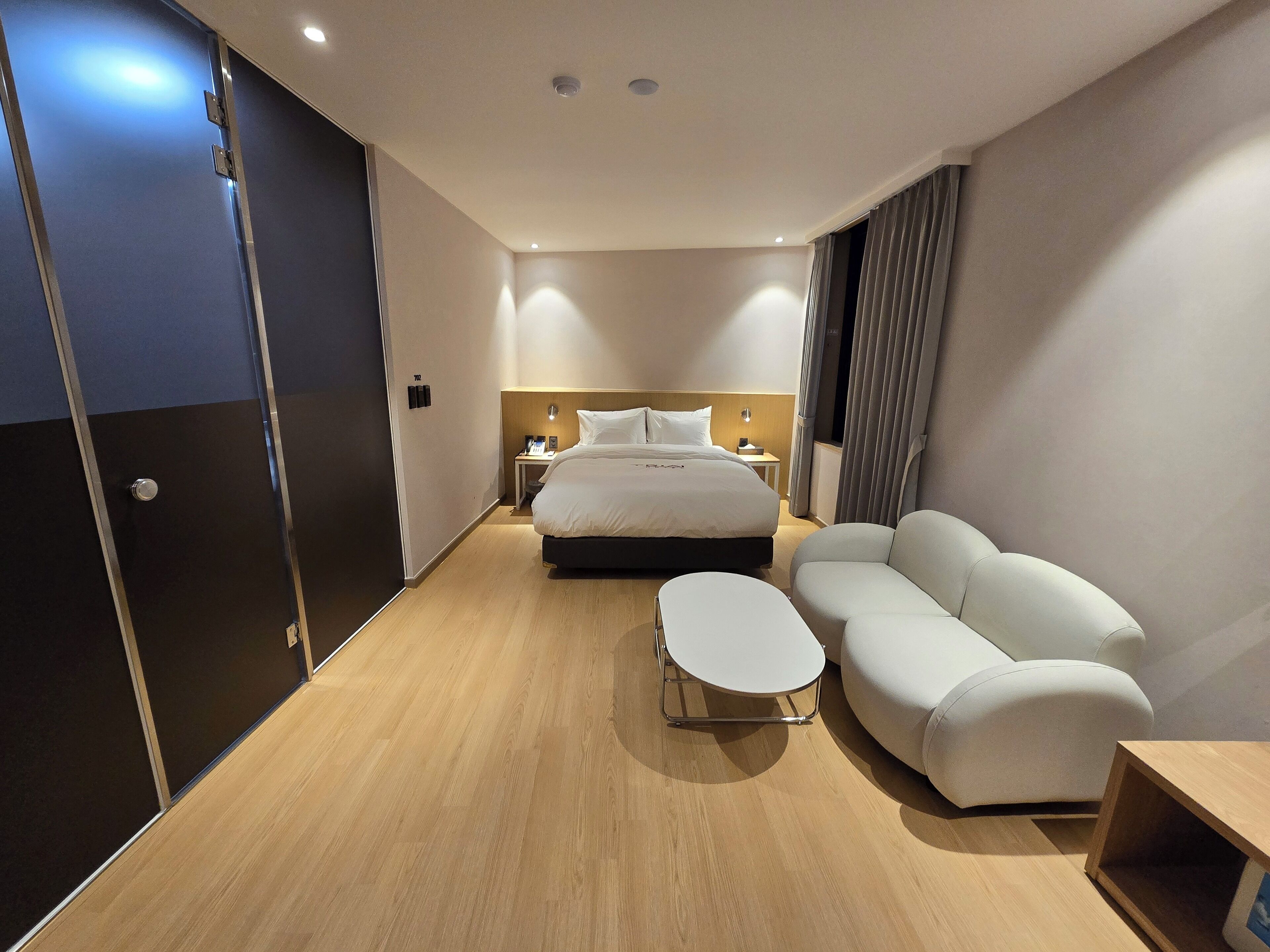 A modern hotel room with a large bed, a sofa, a small table, and a desk with a lamp.