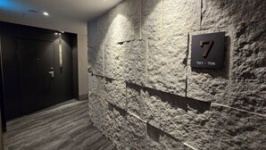 A modern hallway with a textured stone wall, a door with a numeric sign, and a carpeted floor. - Tria Hotel (Seoul)