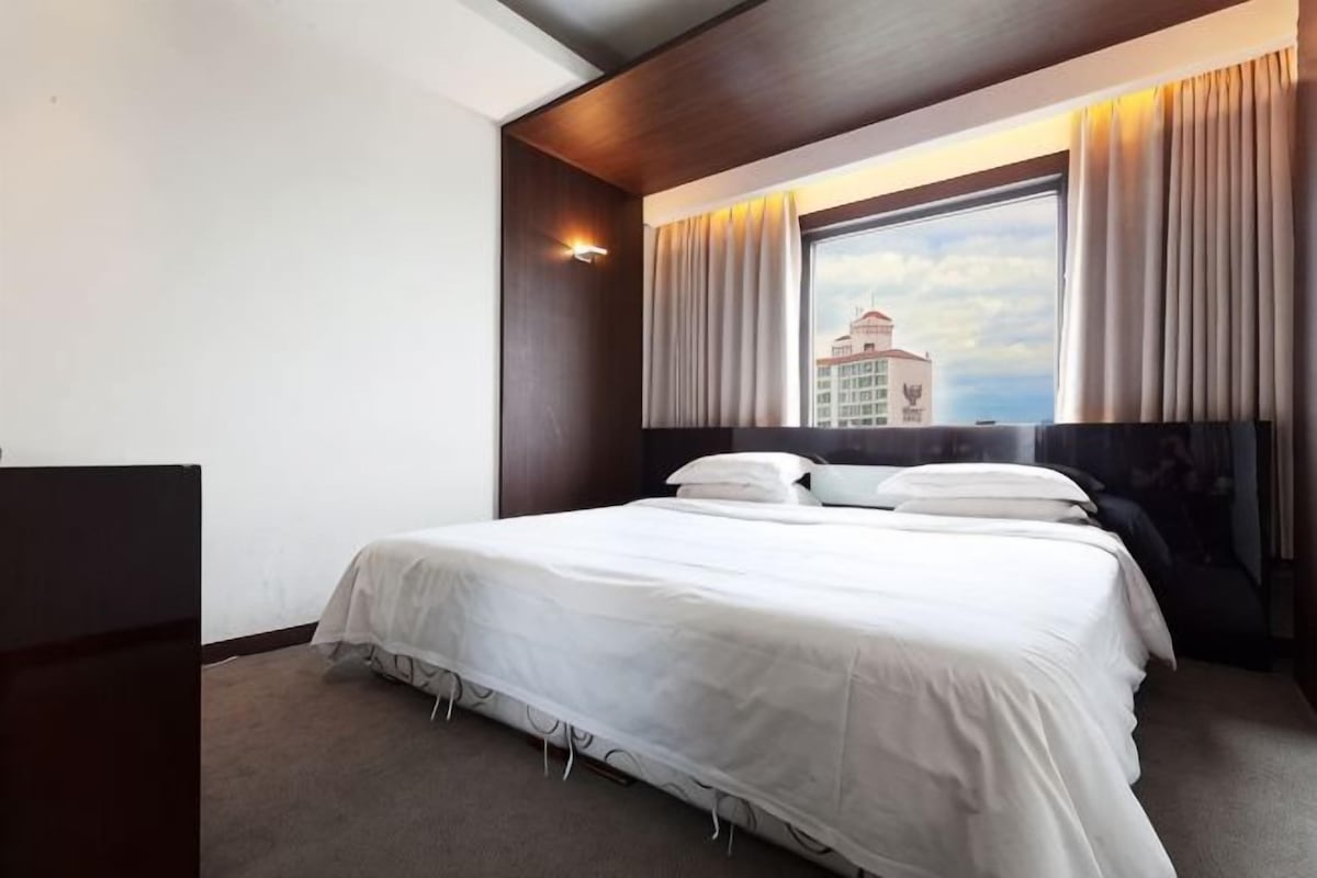 A hotel room with a large bed, a window with curtains, a headboard, and a view of a building outside.