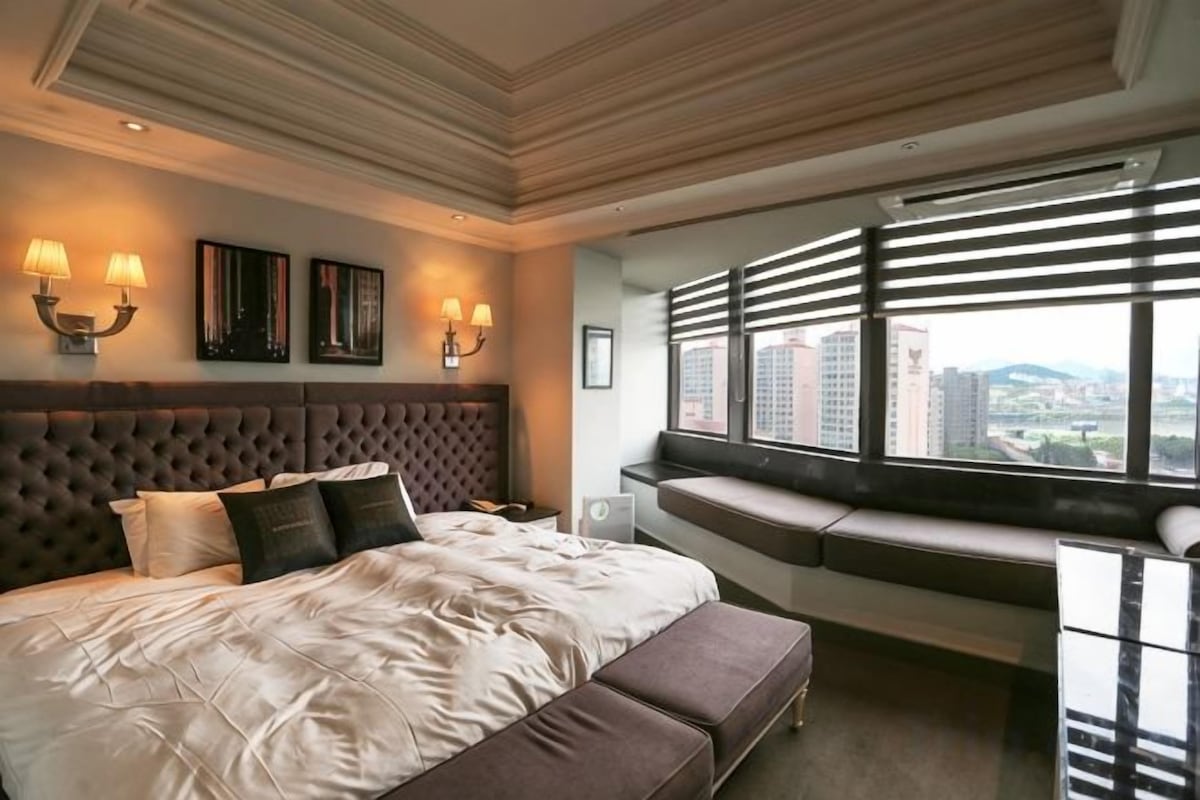 A hotel room with a large bed, a bench, a view of the city, and a window with blinds.