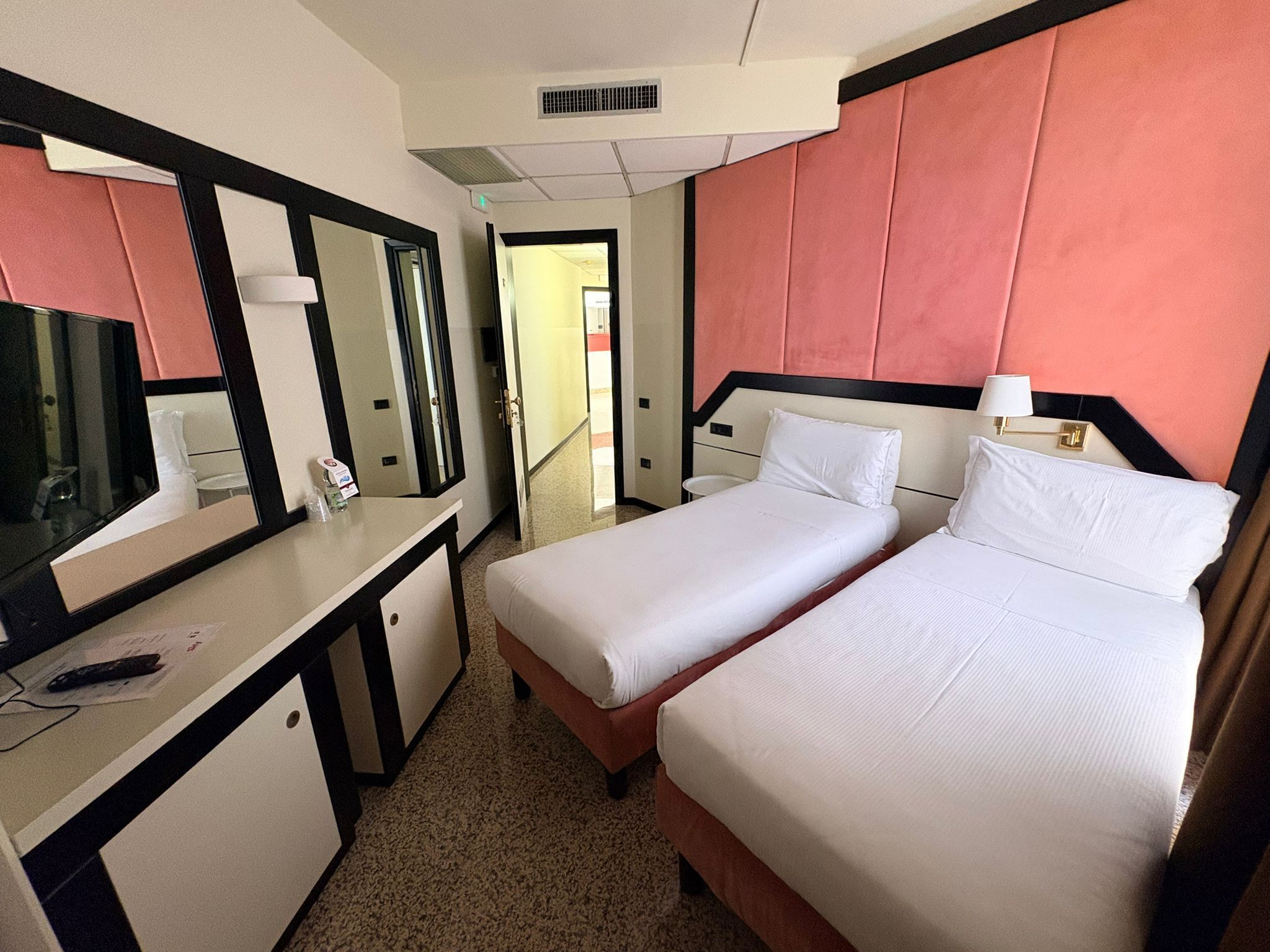 A hotel room with a bed, a desk, a mirror, and a door leading to another room.