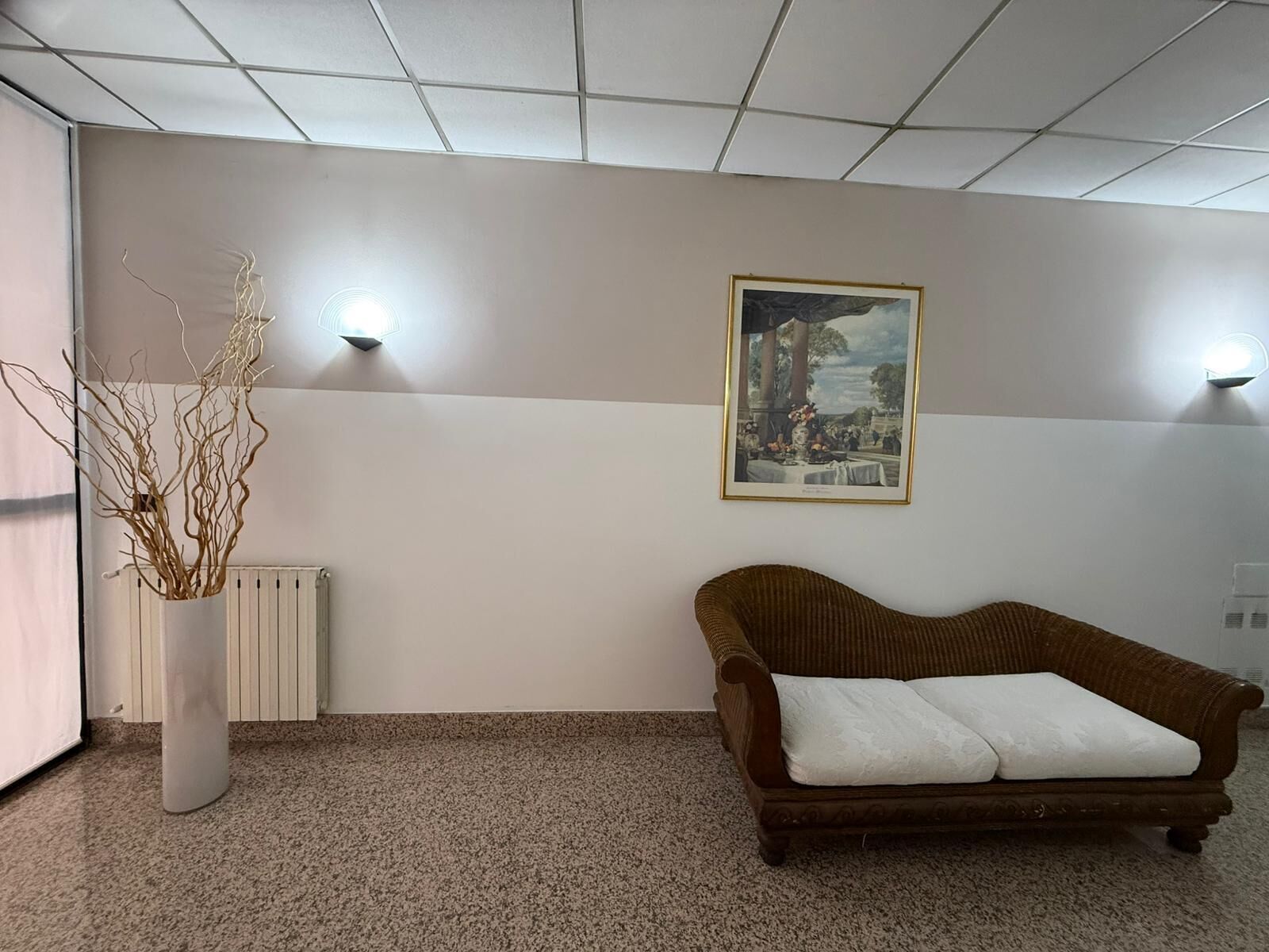 Una habitación con un sofá de mimbre marrón, un radiador blanco, un jarrón blanco con ramas y un cuadro enmarcado en la pared.