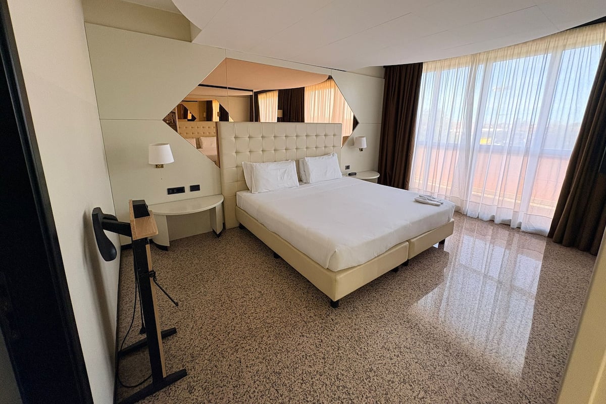 A hotel room with a large bed, a desk, and a chair. There is a window with curtains and a view of the outside.