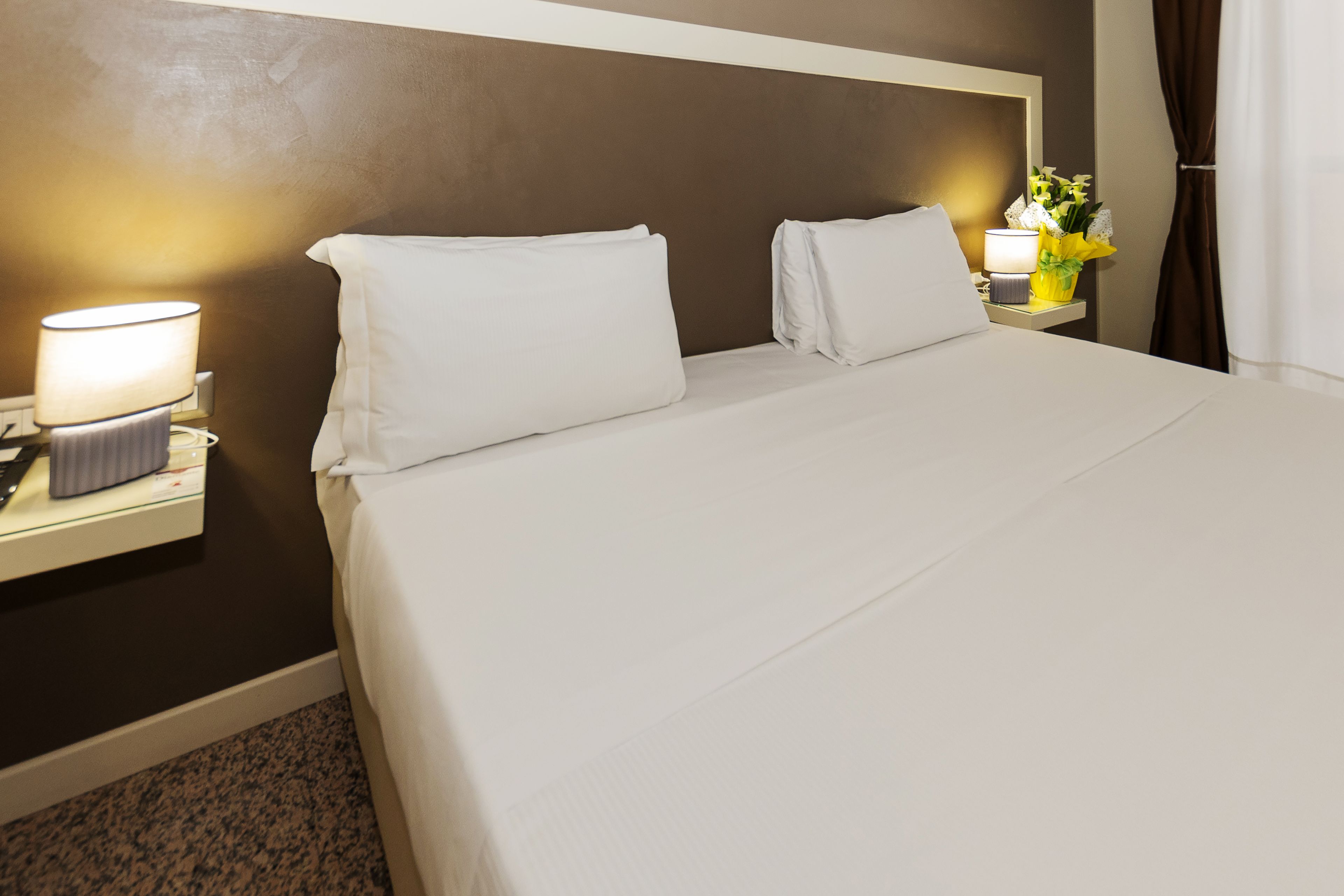 Una habitación de hotel con una cama bien hecha, una lámpara de noche y una mesita de noche con lámpara y un jarrón de flores.