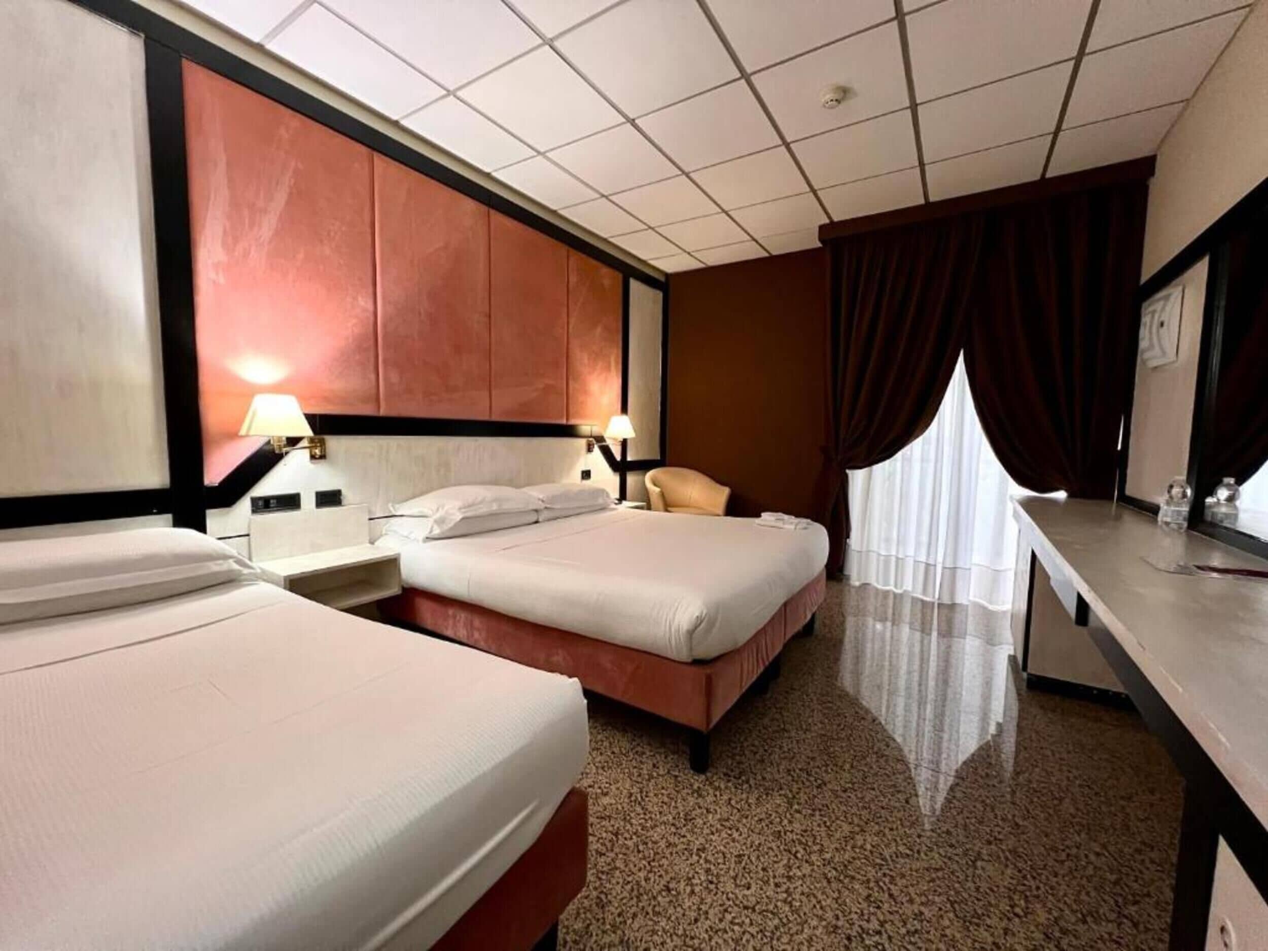 A hotel room with two beds, a desk, a chair, and a large window with curtains.