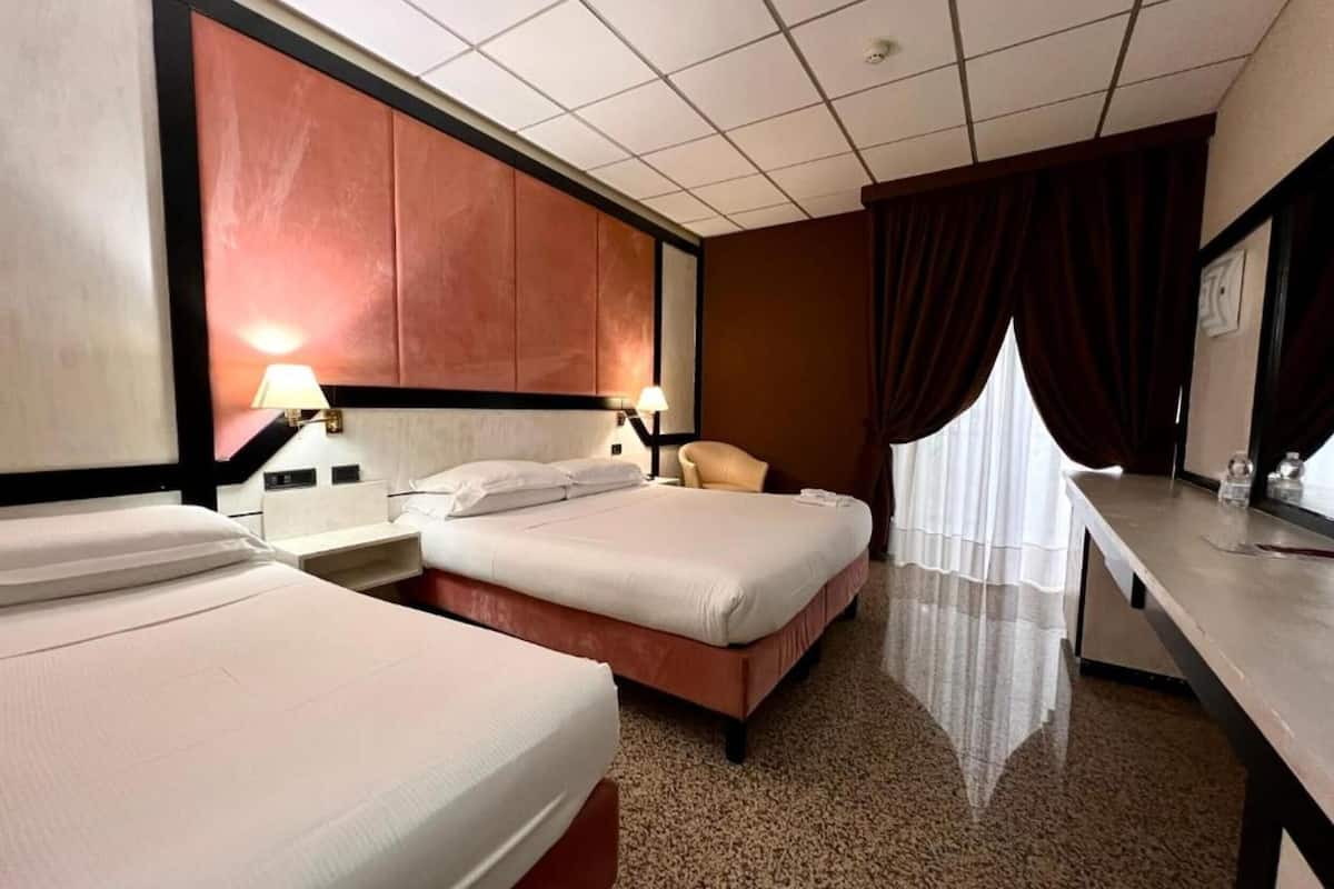 A hotel room with two beds, a desk, a chair, and a large window with curtains.