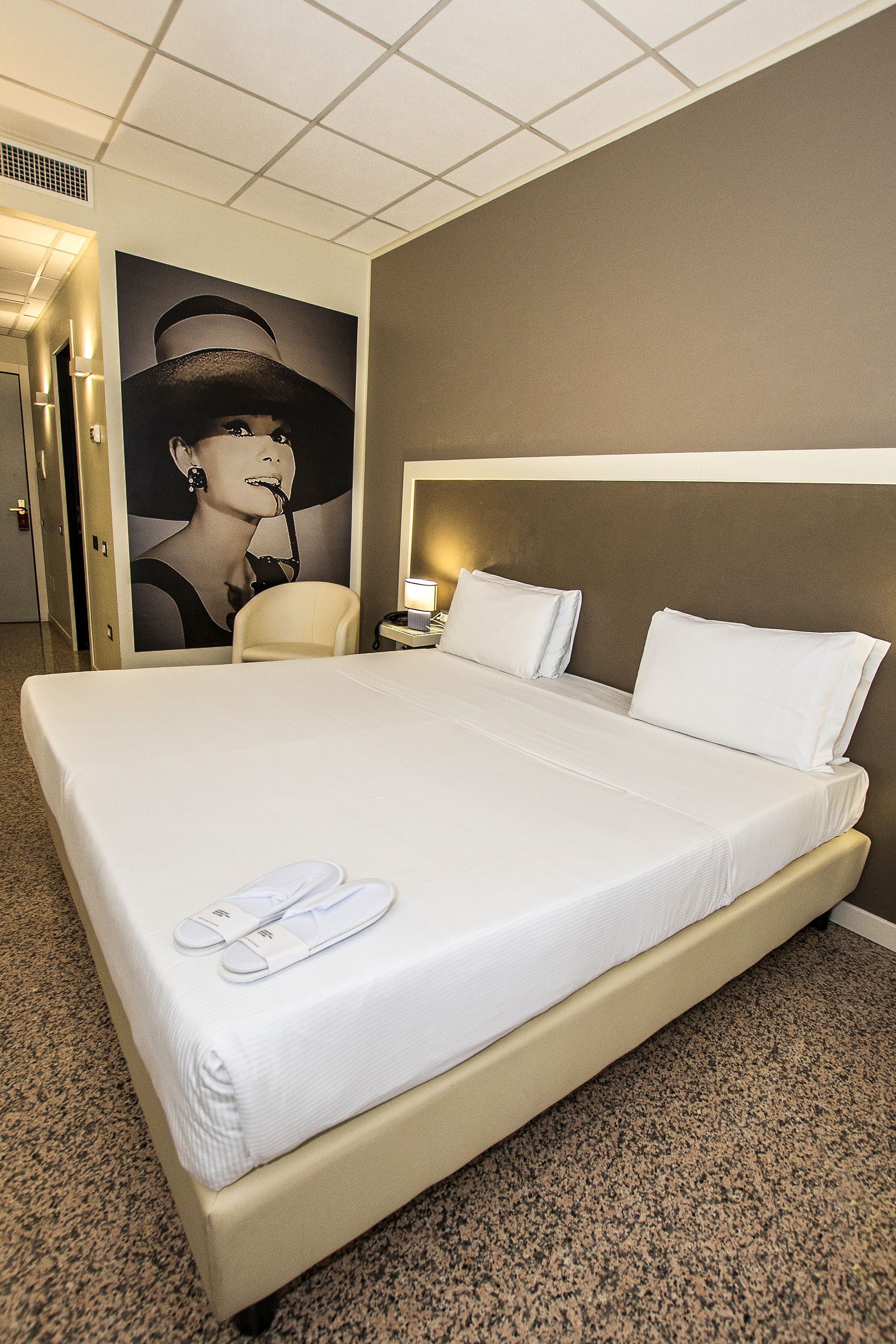 A hotel room with a large bed, a headboard, a nightstand, a lamp, and a poster on the wall.