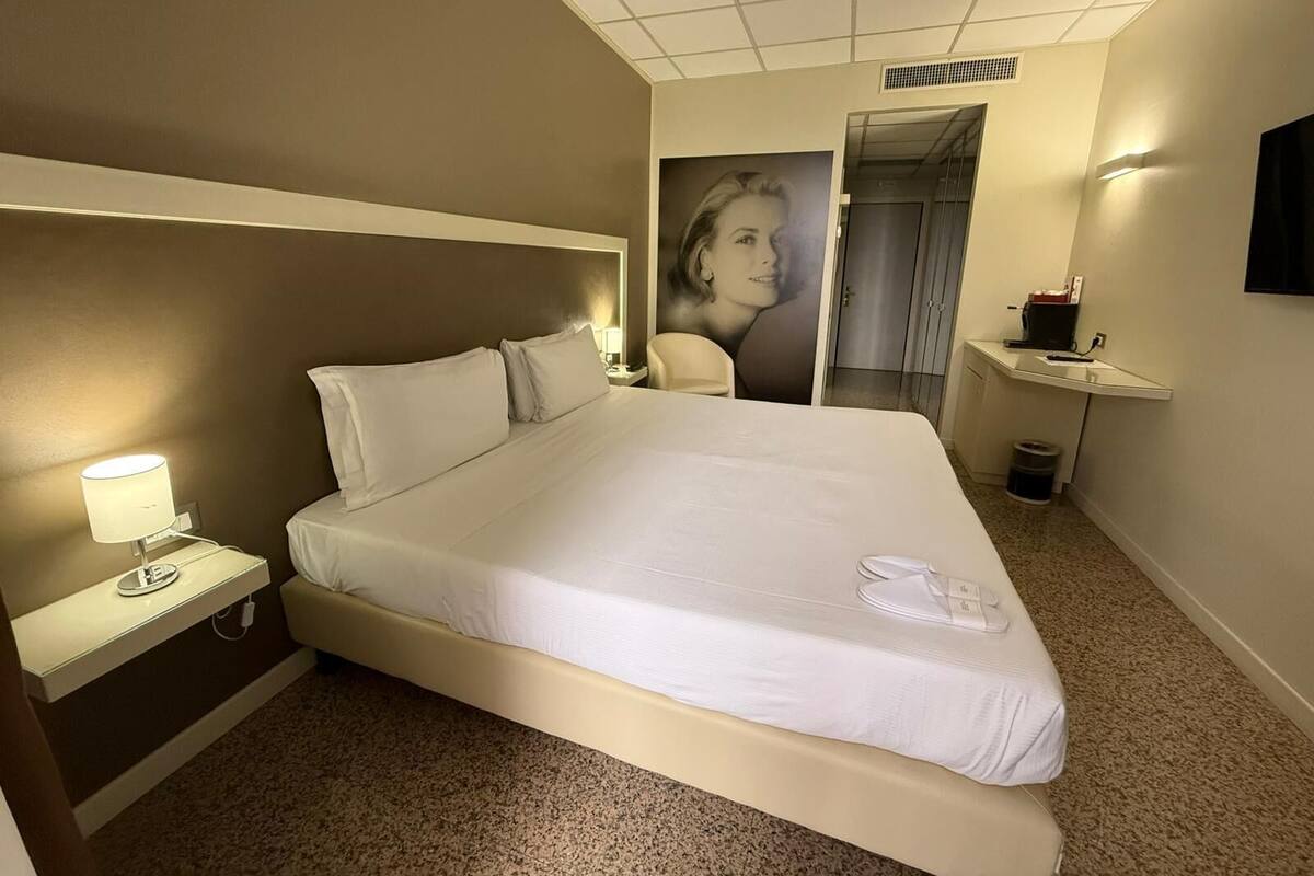 A hotel room with a large bed, a bedside lamp, a small table, a chair, a poster on the wall, and a bathroom door.