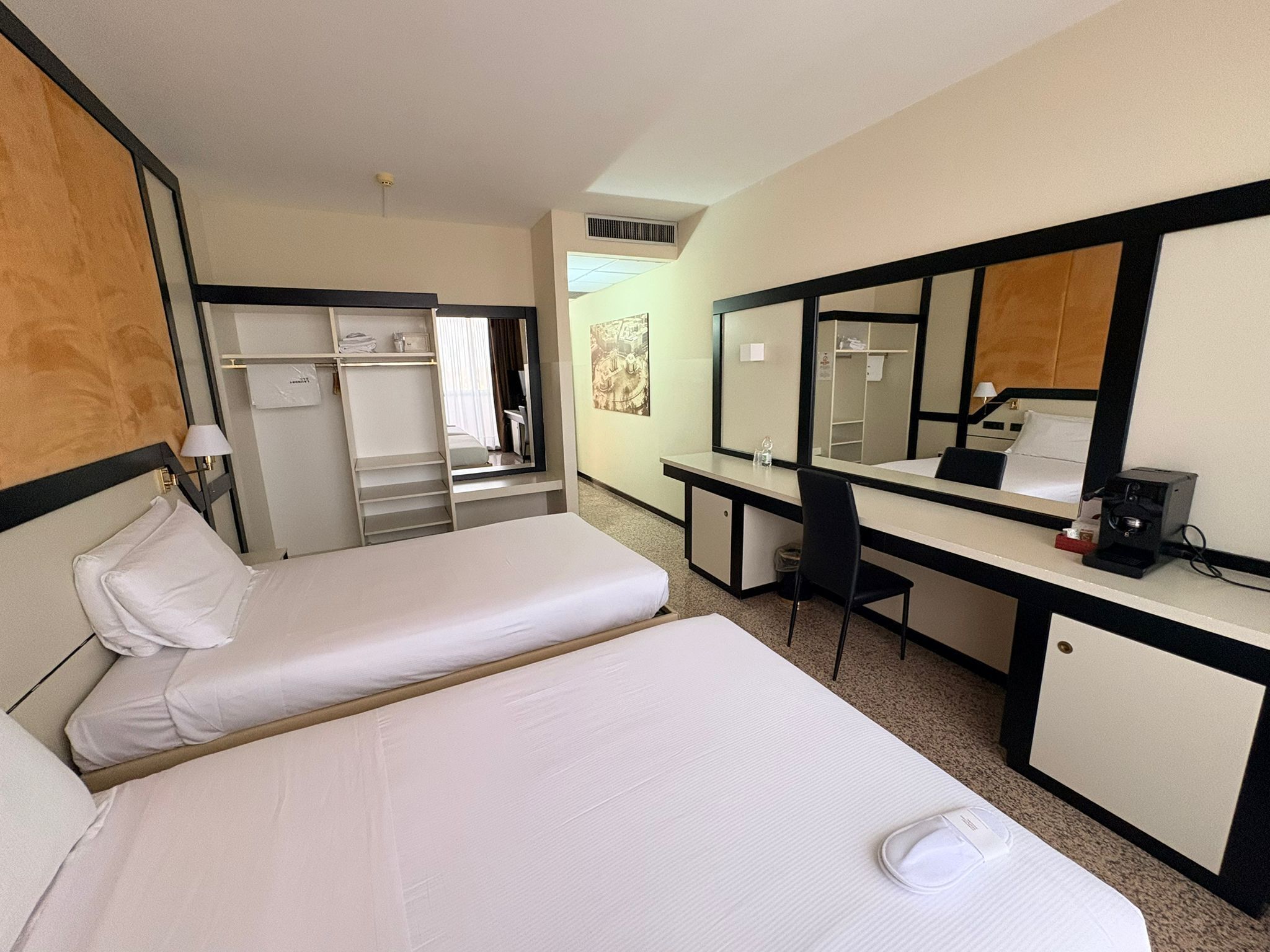 A hotel room with two beds, a desk, a chair, a mirror, and a coffee machine.