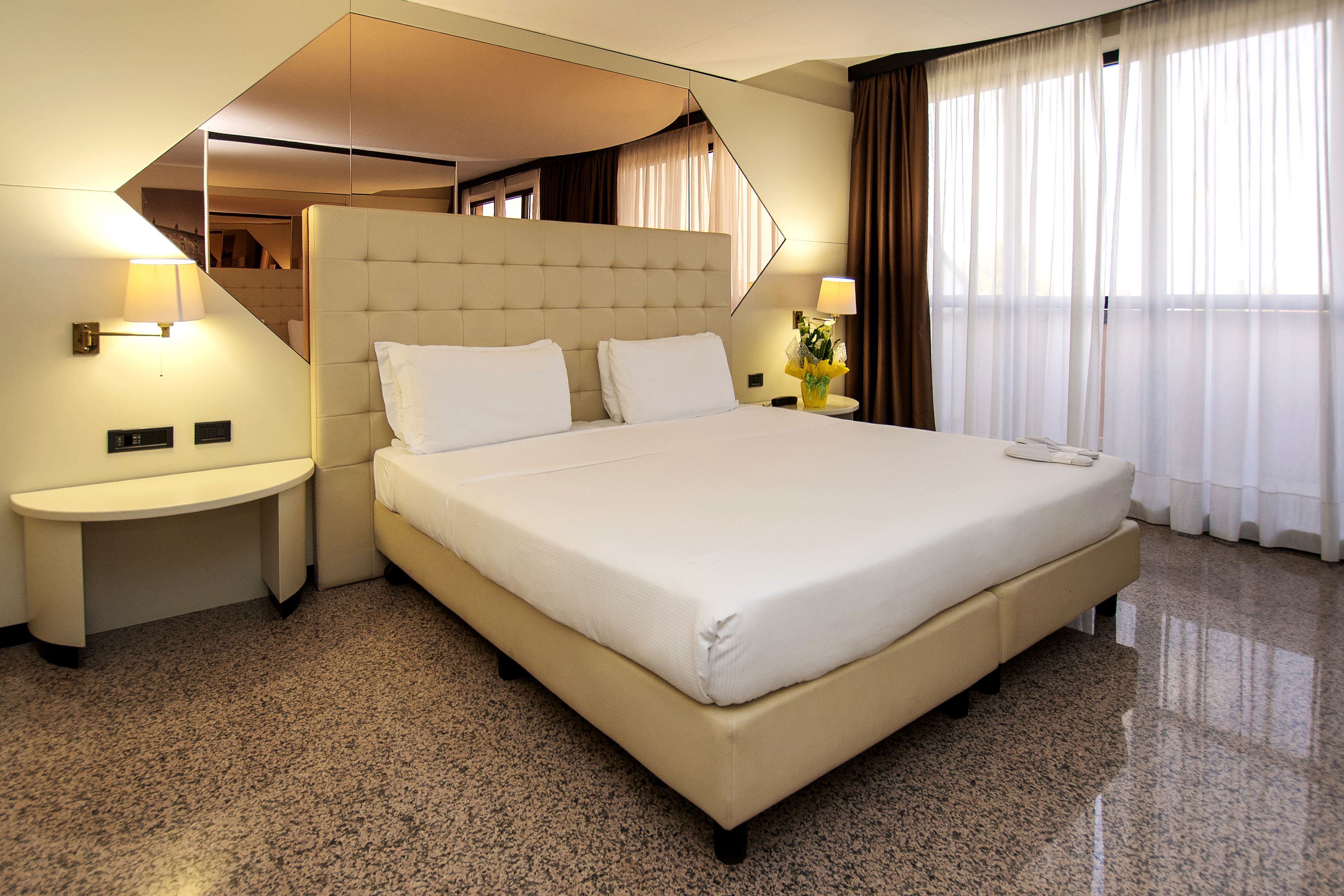 A hotel room with a large bed, two bedside tables, a nightstand, a lamp, and a view of the outdoors through the window.