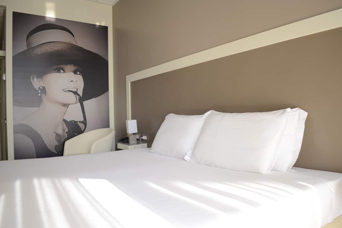 A hotel room with a bed, white bedding, a nightstand with a lamp, and a poster on the wall.