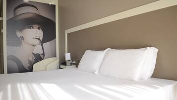 A hotel room with a bed, white bedding, a nightstand with a lamp, and a poster on the wall.