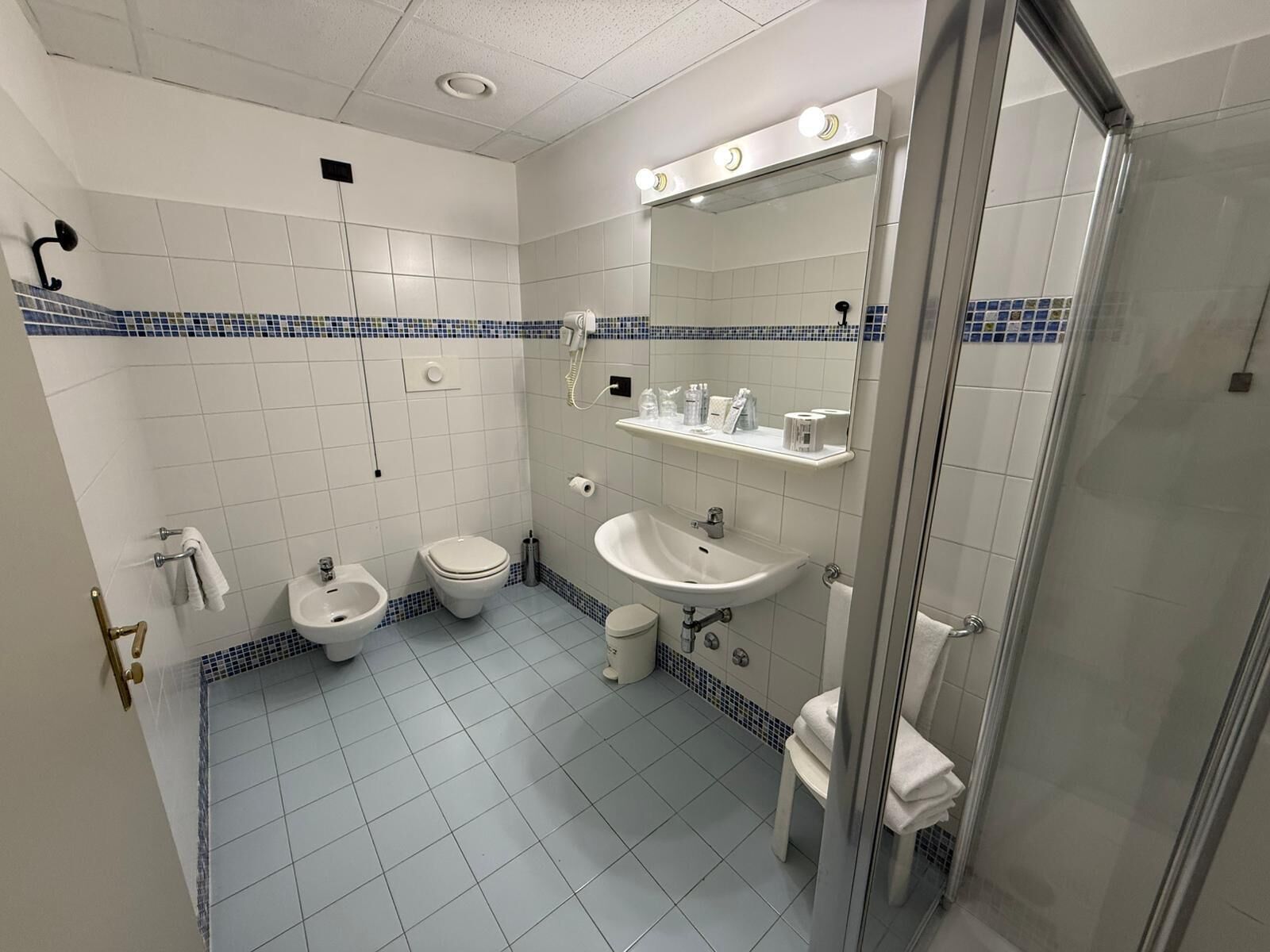 A small, clean bathroom with a toilet, bidet, sink, and mirror. There are shelves above the sink holding toiletries.