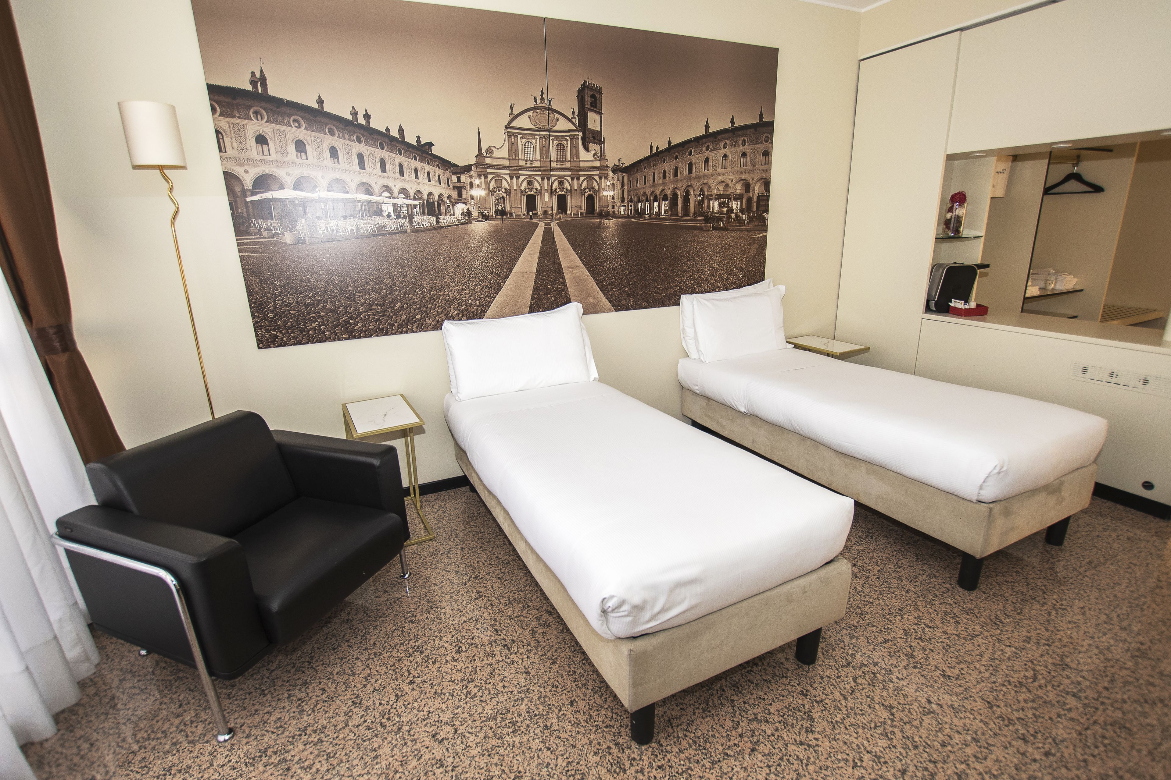 A hotel room with two single beds, a black armchair, a small table, a mirror, and a large cityscape painting on the wall.