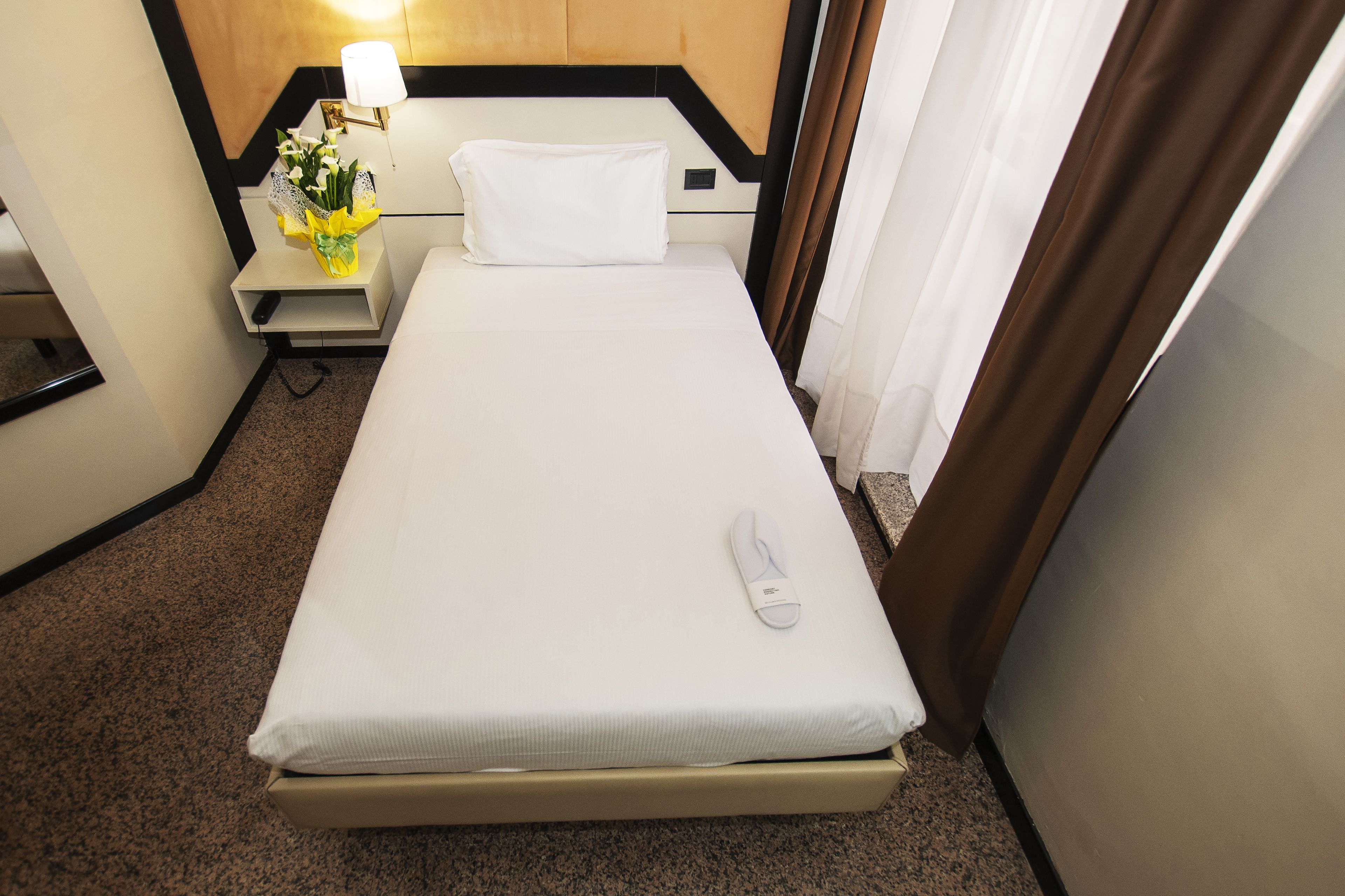 A hotel room with a single bed, a nightstand with a lamp, and a small table with a vase of flowers.