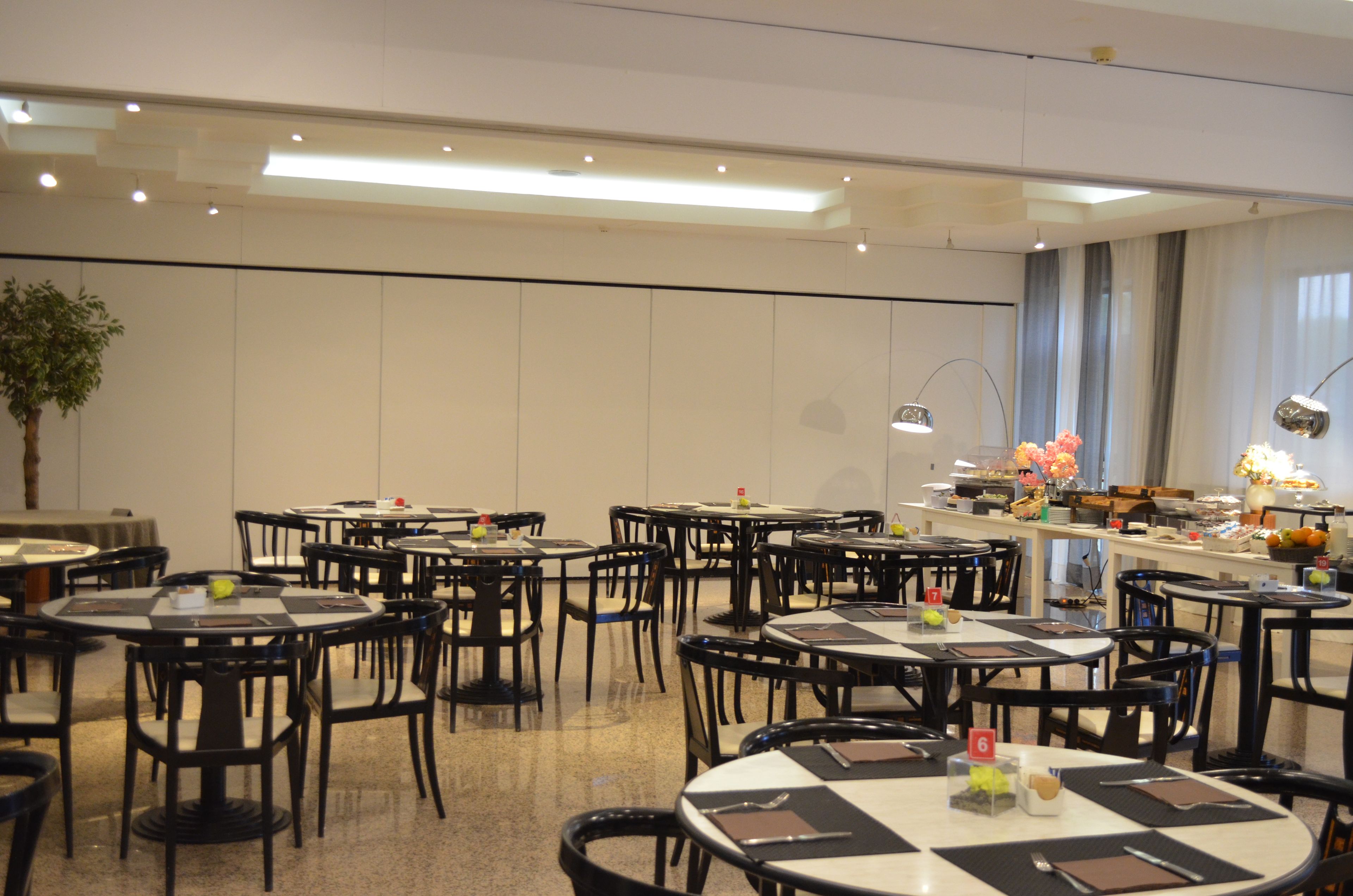 Une salle à manger d’hôtel avec plusieurs tables rondes et des chaises, un buffet et une grande plante en pot.