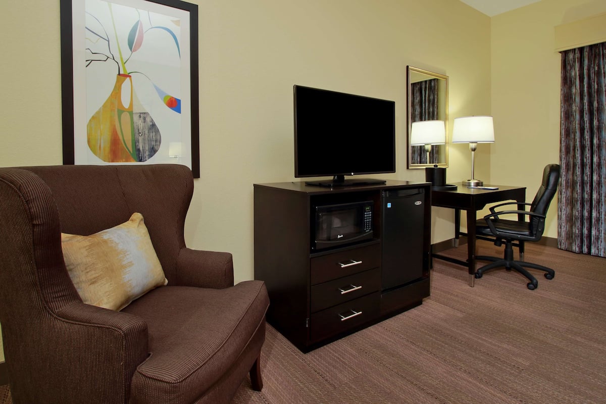 Quarto de hotel com poltrona marrom, televisão de tela plana, micro-ondas, escrivaninha com luminária e uma cadeira.