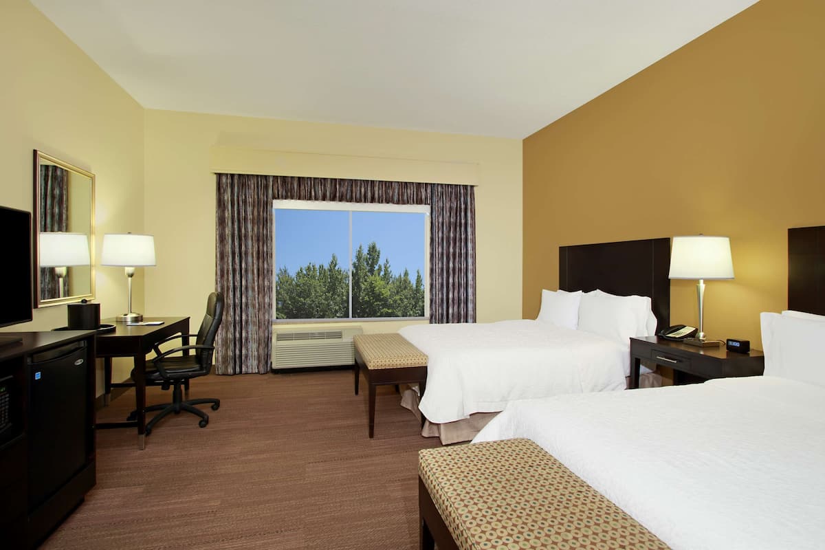 Quarto de hotel com duas camas, uma secretária com cadeira, televisão e janela com cortinas.