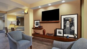 Un hall de hotel con dos sillones, un televisor de pantalla plana montado en la pared, un cuadro con un coche antiguo y un pequeño reloj en la repisa.