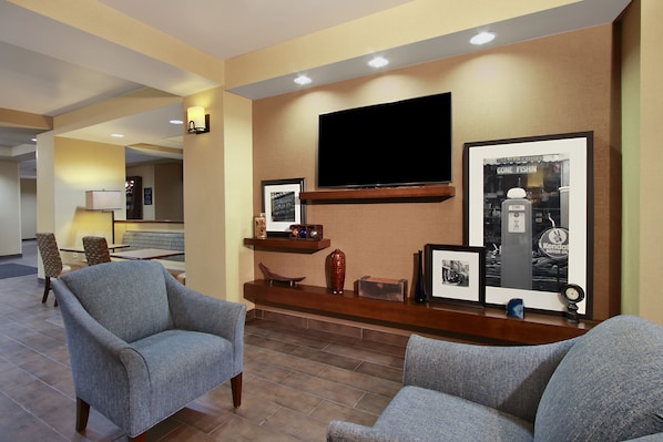 Un hall de hotel con dos sillones, un televisor de pantalla plana montado en la pared, un cuadro con un coche antiguo y un pequeño reloj en la repisa.