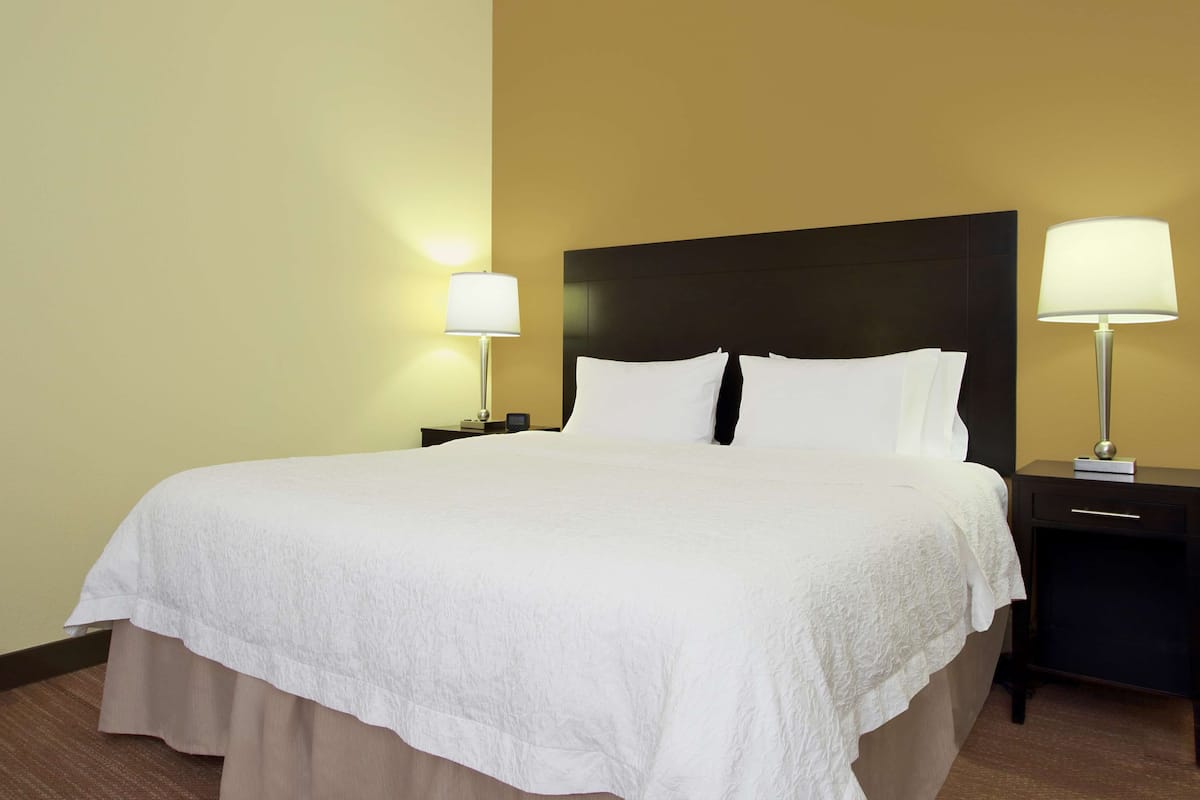 Cama bem arrumada com colcha e almofadas brancas, com duas mesas de cabeceira com luminárias, em um quarto com parede lisa e piso coberto de carpete.