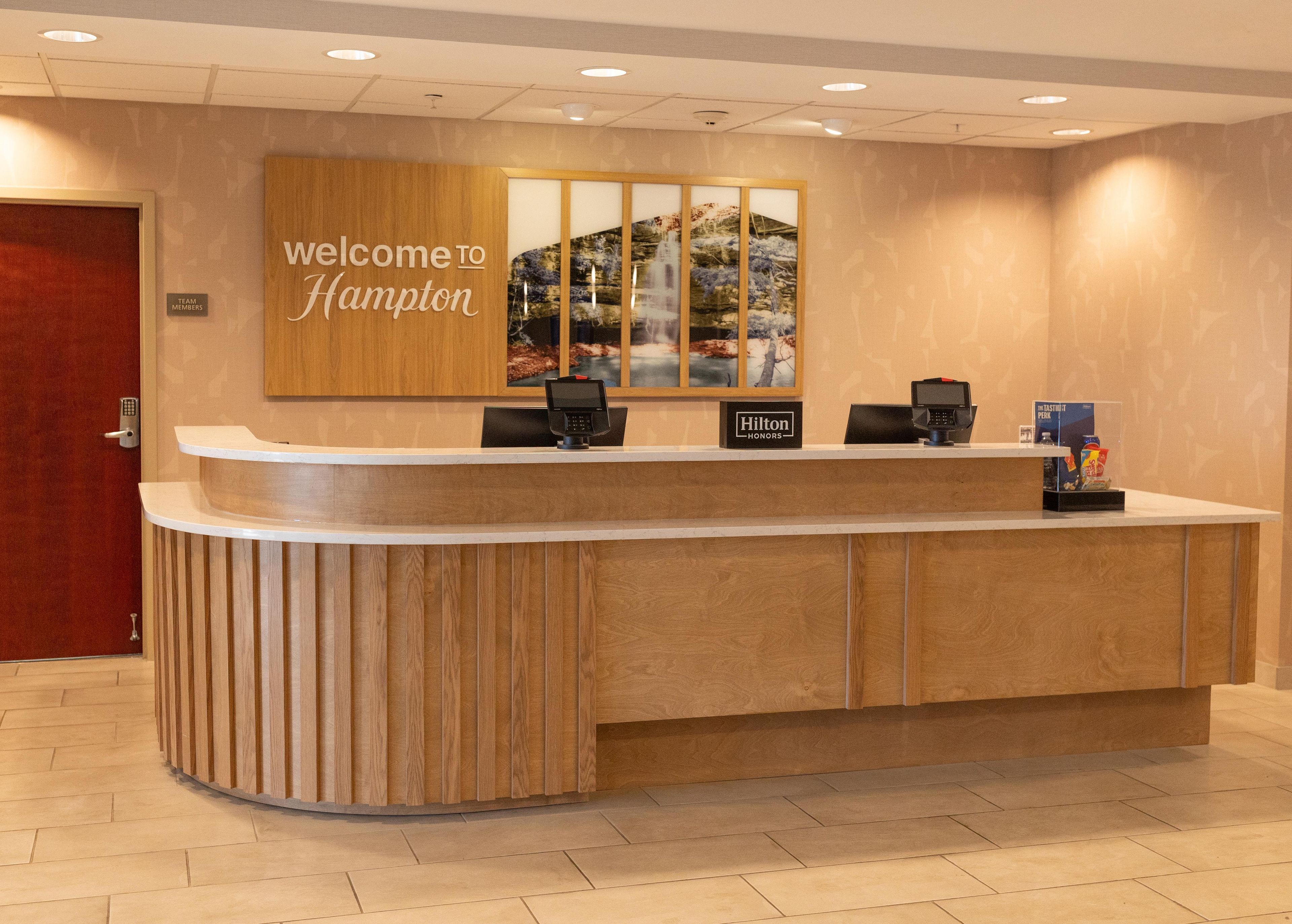 A reception desk with a 'Welcome to Hampton' sign, a 'Hilton Honors' badge, and a digital kiosk.