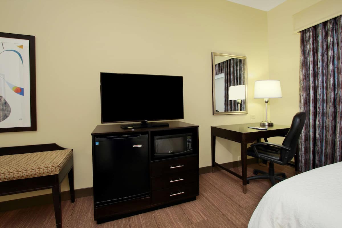 Quarto de hotel com cama, escrivaninha com cadeira, televisão, micro-ondas e frigorífico.