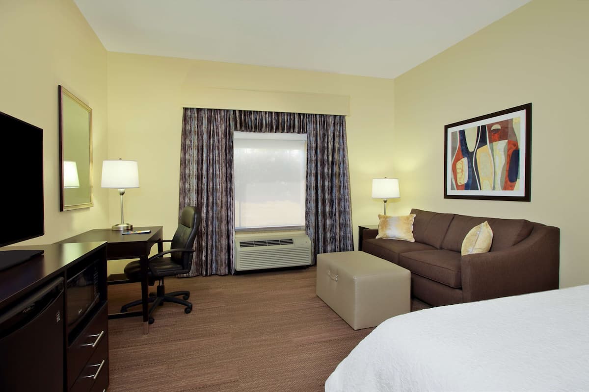 Quarto de hotel com cama, escrivaninha com computador, cadeira, televisão, janela com cortinas, ar condicionado e um quadro na parede.