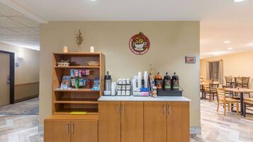 Zona de recepción de hotel con un mostrador de madera, una máquina de café y un cartel anunciando el desayuno.