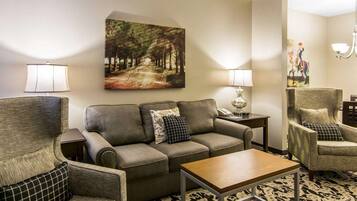 A living room with a sofa, armchairs, a coffee table, a painting of a forest path, and a lamp.