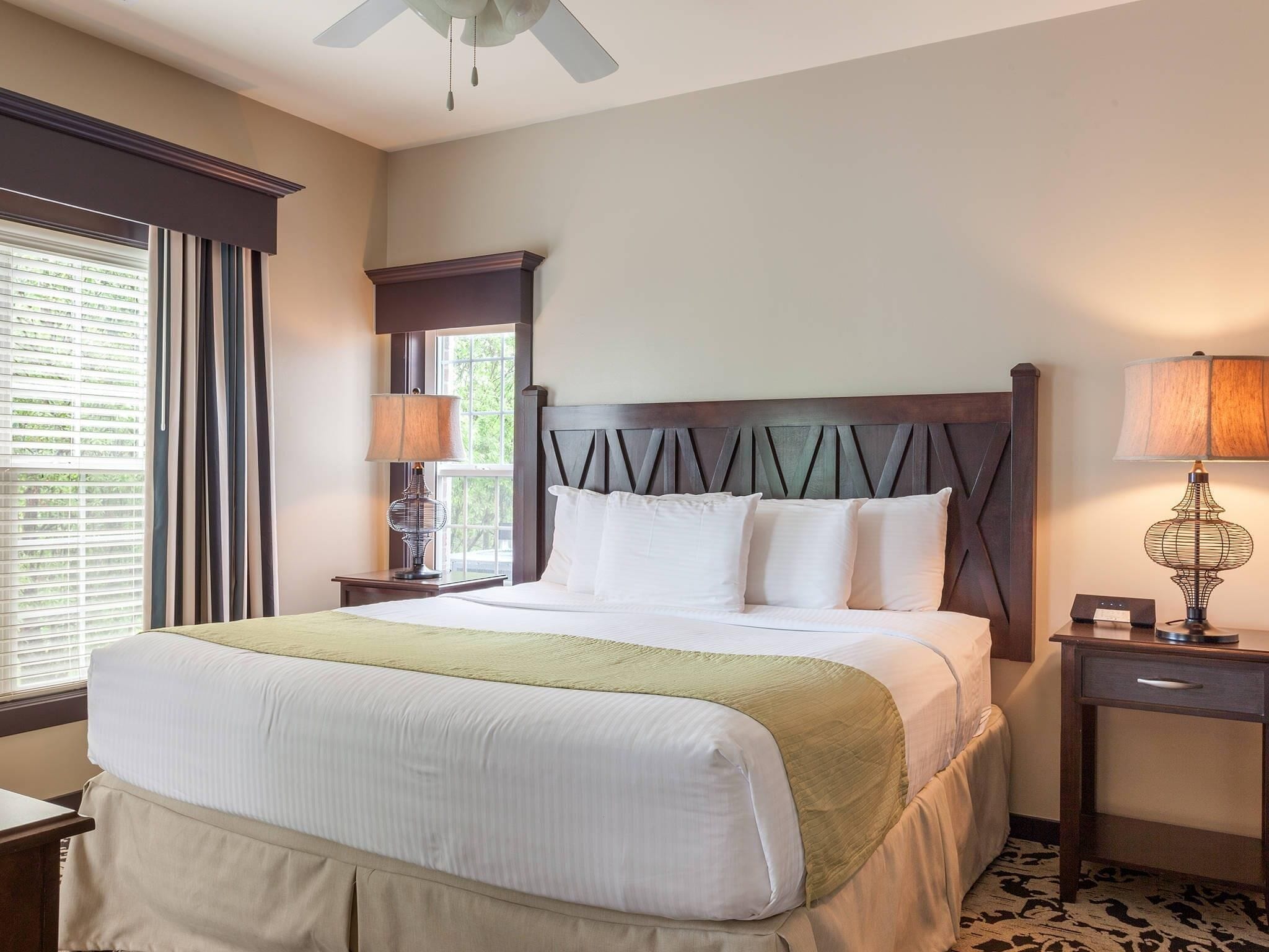A hotel room with a large bed, two bedside tables with lamps, a window with curtains, and a ceiling fan.