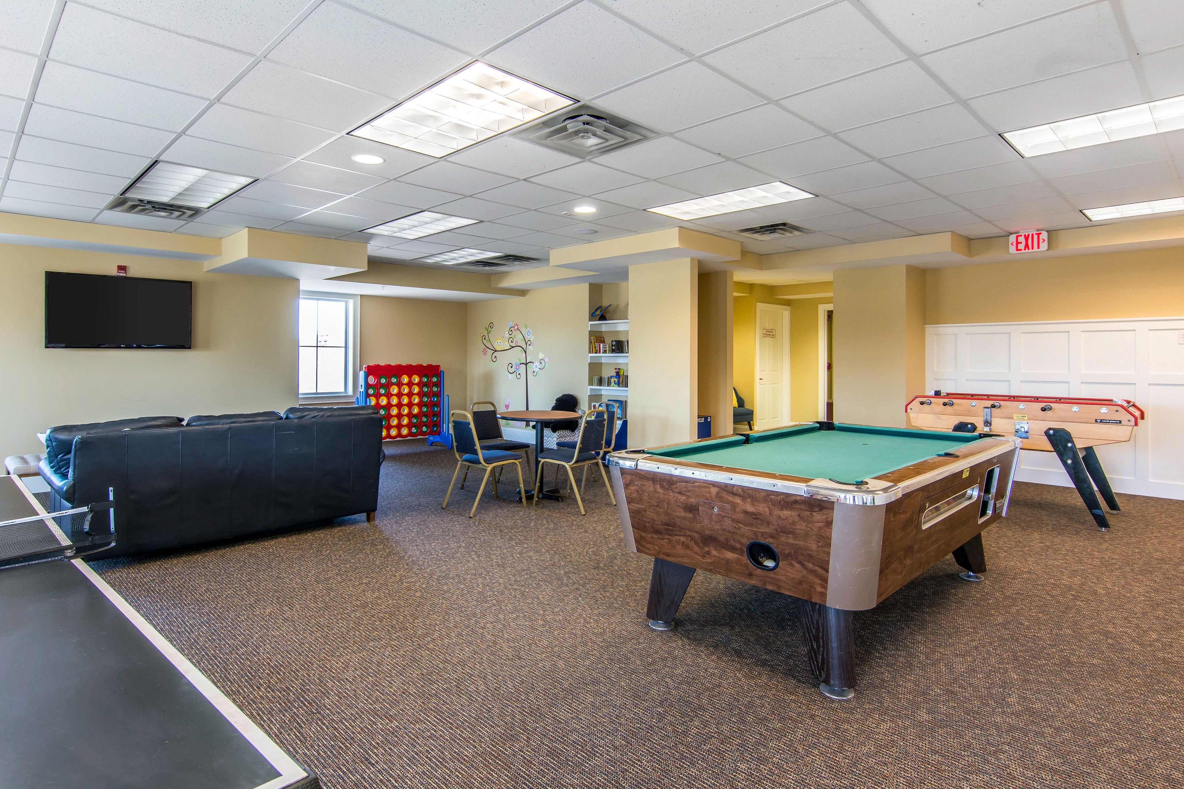 A pool table, a foosball table, and a billiards table in a recreational room with a carpeted floor and ceiling lights.