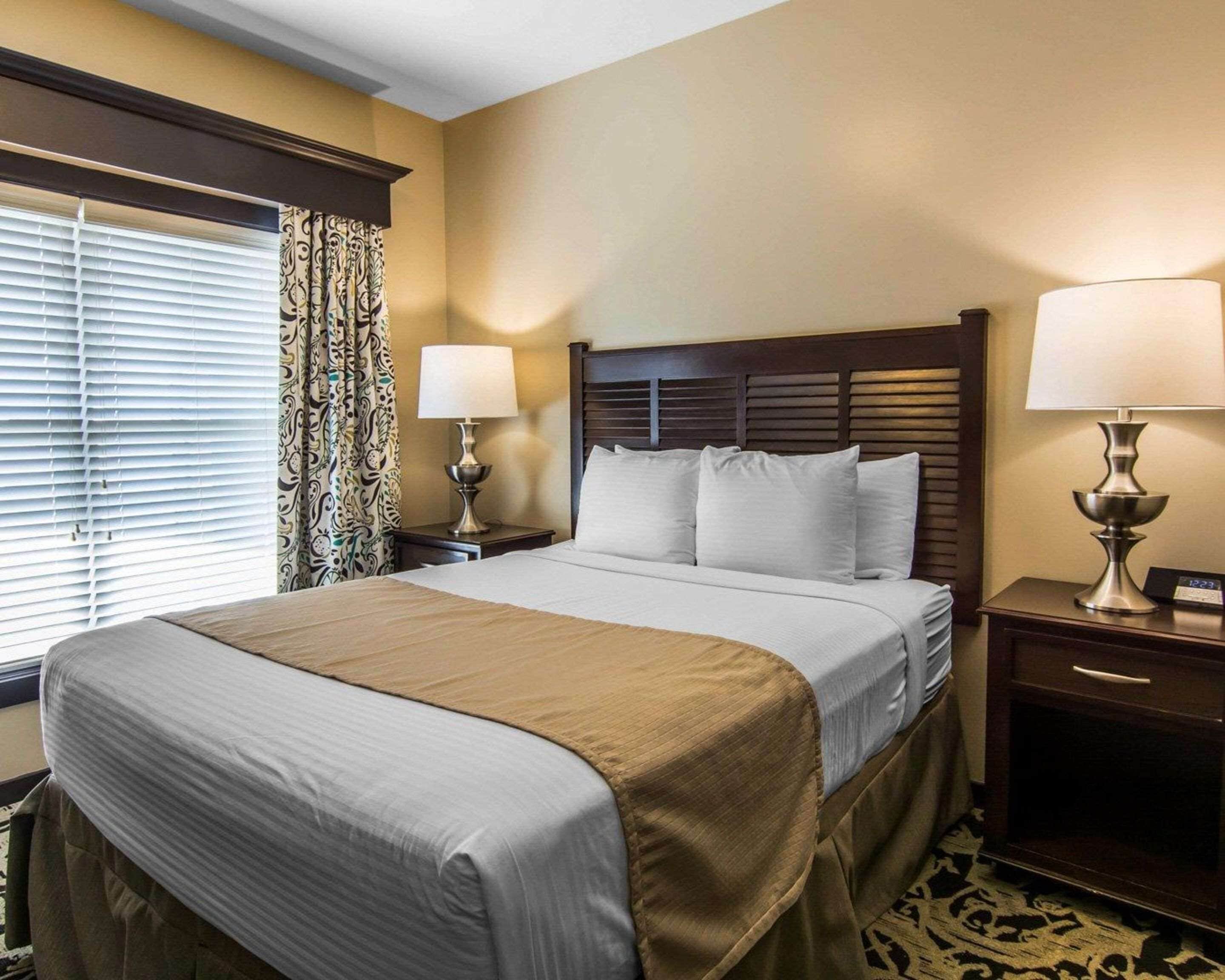 A hotel room with a large bed, two bedside tables with lamps, a window with blinds, and a patterned curtain.